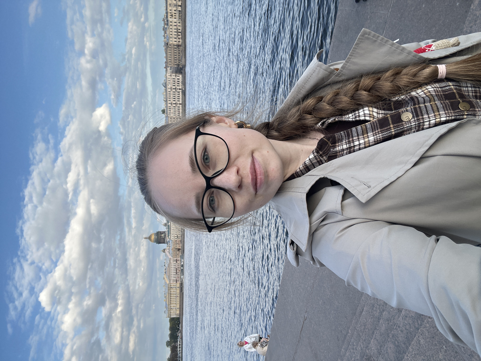 A selfie shows a woman with long, braided hair wearing glasses and a tan trench coat over a plaid shirt. In the background, across a body of water, is the cityscape of Saint Petersburg, Russia. The distinct gold dome of St. Isaac's Cathedral is visible among the buildings. To the left, the sky is blue with scattered clouds. In the lower background, two pedestrians are visible. The woman in the selfie is standing on a stone embankment.
