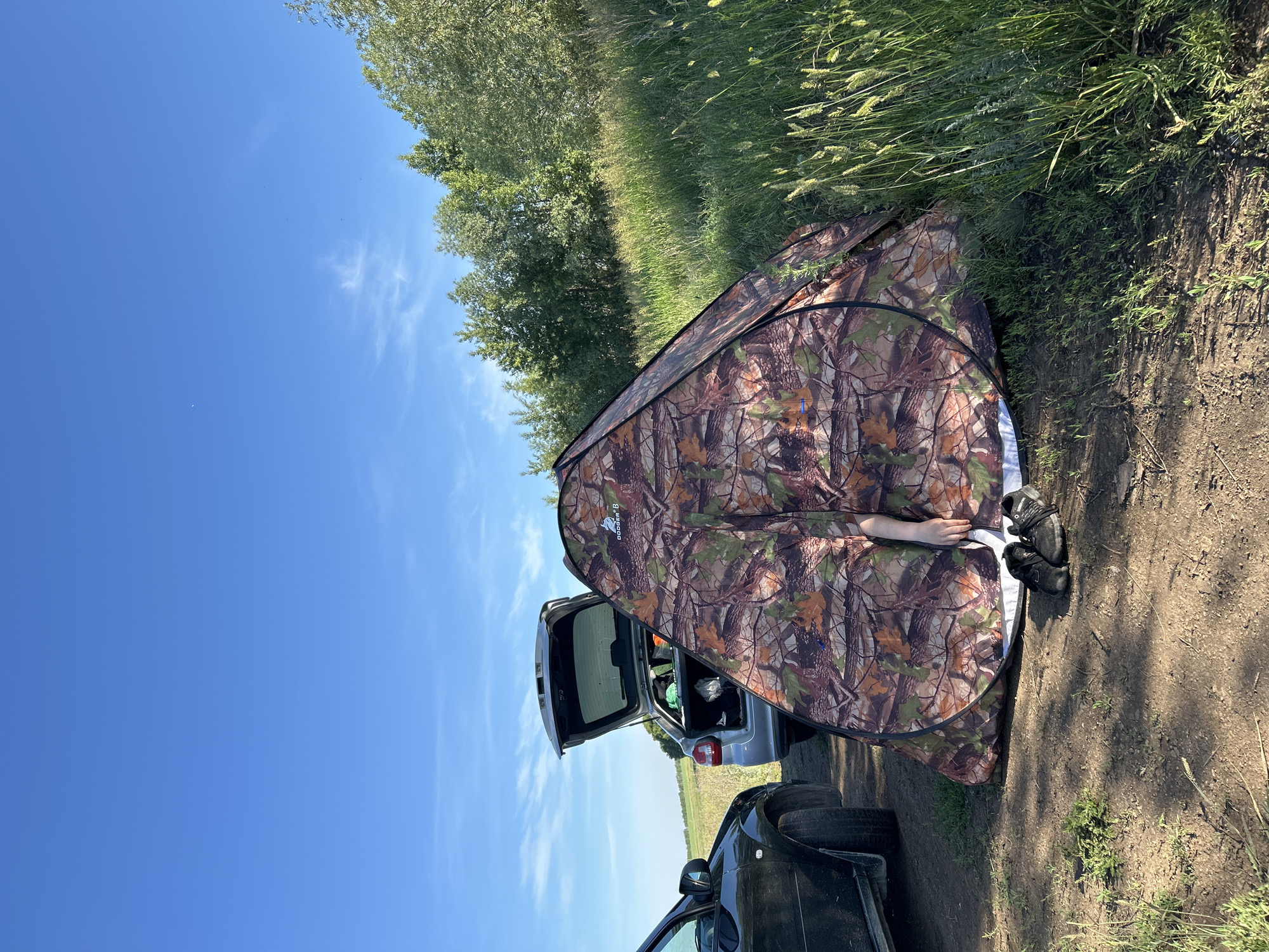 The image depicts a campsite with a camouflage tent set up in a grassy field under a clear blue sky. Two cars are parked nearby; one is a dark-colored car on the left, and a silver hatchback with its trunk open is visible to the left of the tent. The trunk of the silver car contains some items. A person's arm, draped over the edge of the tent, is visible. A pair of black shoes rests on the ground in front of the tent. The background consists of a line of trees and a field of tall grass. The scene appears to be located in Matraevo, Russia.
