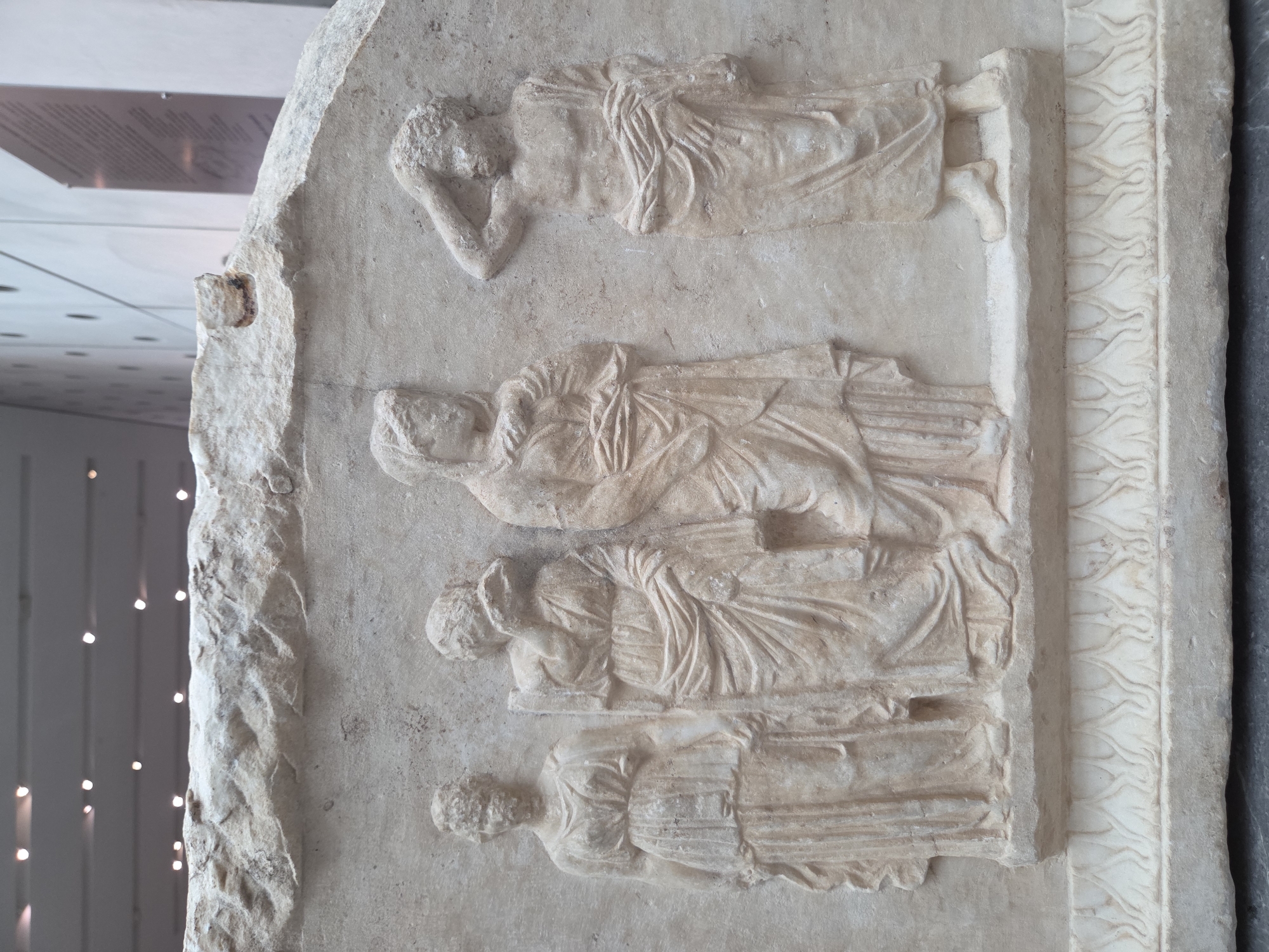 This is a detailed photograph of an ancient Greek relief carving, likely from a stele or sarcophagus, displayed in the Acropolis Museum in Athens, Greece. The relief depicts four figures in profile or partial profile, carved in marble.
From left to right:
The first figure is a standing female, wearing a chiton with a belted waist and draping sleeves, and a peplos over it. Her hair is styled in a bun.
The second figure, also female, stands slightly behind and to the right of the first. She is also draped in a chiton and peplos, with her right hand raised to her face, obscuring her mouth. Her hair is styled similarly.
The third figure, a female, is positioned to the right of the second. She wears a chiton and peplos. Her left arm is bent across her chest, and her hands are clasped or holding drapery. Her head is turned to her left.
The fourth figure, a male, stands to the far right. He is bare-chested, with his right arm raised to his forehead, hand resting on his head. He wears a draped garment around his waist and hips. His hair is curly.
The figures are carved in low relief on a textured background. They stand on a raised platform with a decorative border at the base, featuring a pattern of palmettes or lotus blossoms. The top edge of the relief is broken and uneven. The overall condition suggests significant age and wear. The environment appears to be a museum exhibit, with subtle lighting and a blurred background that hints at other displays or architectural elements.