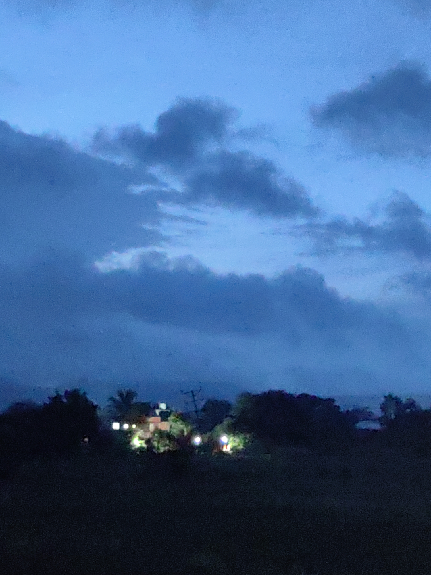 This image depicts a dimly lit landscape at dusk or night, characterized by a dark foreground of vegetation and a background of silhouetted trees and structures. The sky is a deep blue, with scattered, dark clouds obscuring the upper portion of the scene. In the mid-ground, several illuminated lights are visible, suggesting human habitation or activity. These lights are clustered around what appears to be a building or group of buildings, with some of the lights casting a yellowish glow. The overall impression is one of a rural or semi-rural setting, possibly on the outskirts of a town or city. Without further information or context, it is impossible to definitively identify this as the city of Khuldabad, India. However, the presence of vegetation and structures is consistent with a landscape that could be found in that region.