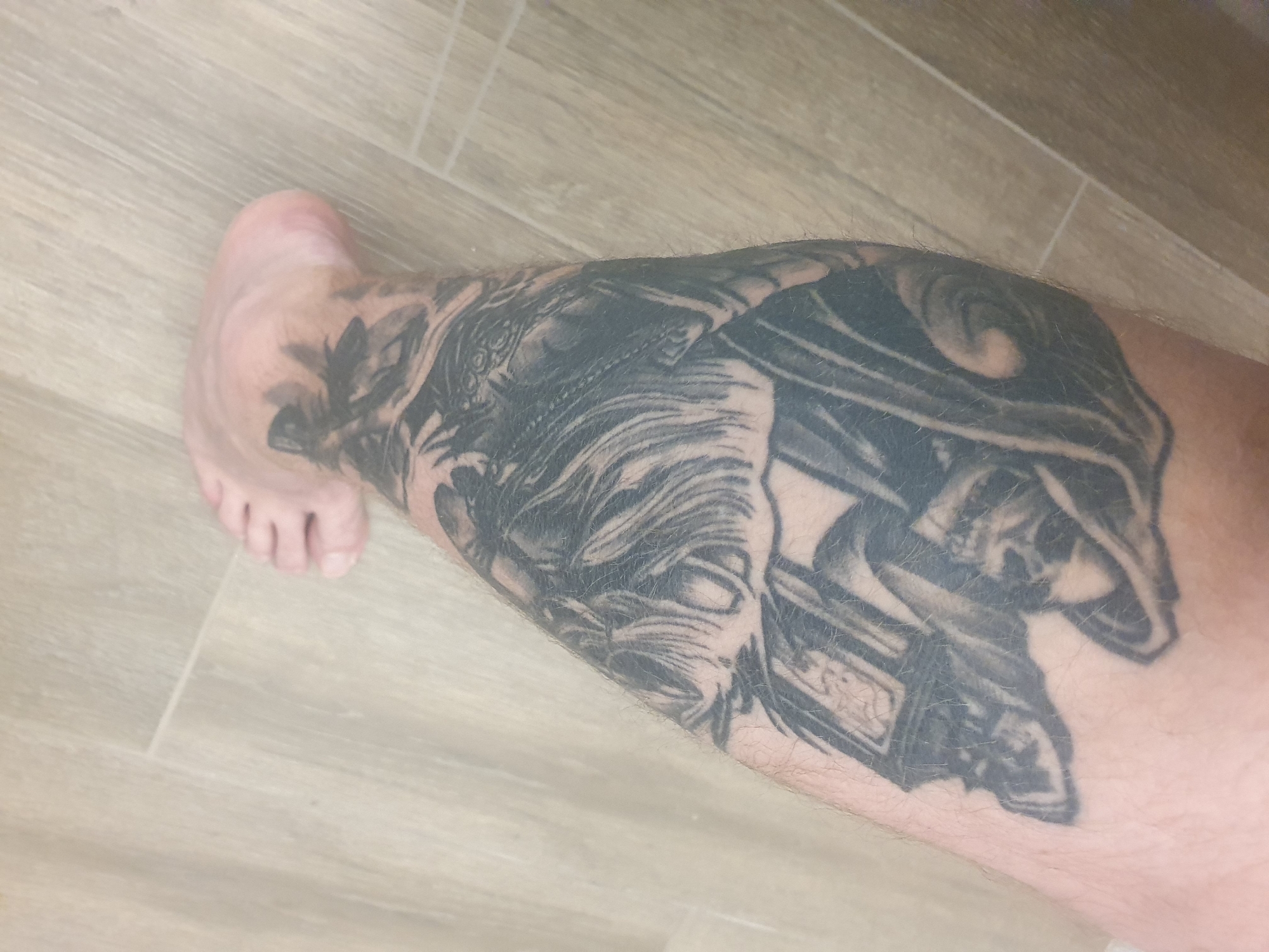 A close-up, indoor shot shows a person's lower leg and foot resting on a light-colored, wood-patterned floor. A large, monochrome tattoo covers the majority of the lower leg. The tattoo's design appears intricate and detailed. The person's foot is visible at the left edge of the frame. The location is unknown and there is no information to determine if this image was taken in Plovdiv, Bulgaria.
