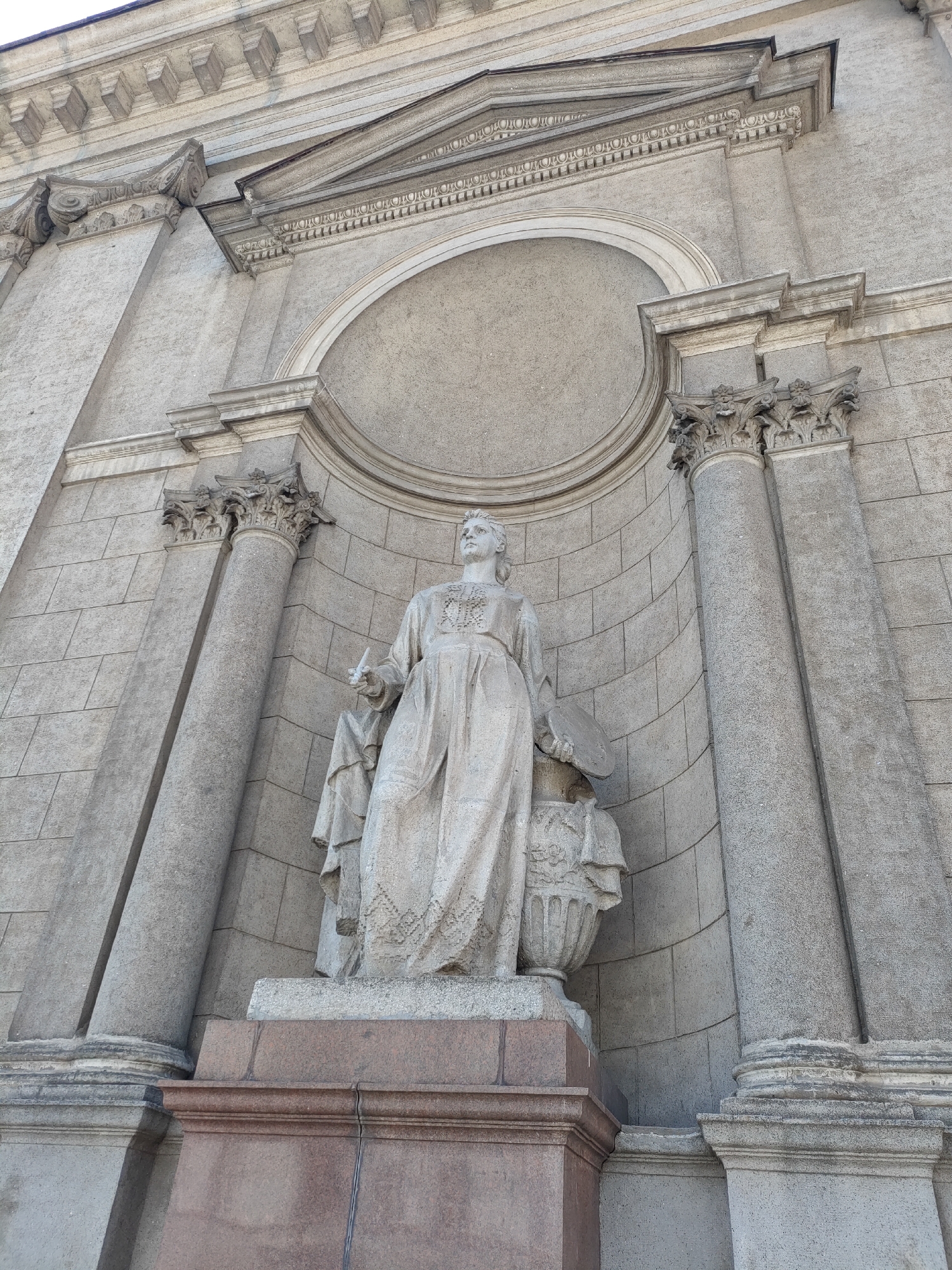 The image shows a low-angle, full-shot of a stone sculpture of a woman positioned in a niche of a building. The sculpture is of a woman wearing a long, flowing robe and holding a palette and brush in her hands. She is standing next to a large urn. The building behind her is made of stone and features classical architectural elements, including columns and ornate carvings. The overall impression is one of grandeur and artistic expression.
This sculpture is located on the facade of the Belarusian State Academy of Arts in Minsk, Belarus.