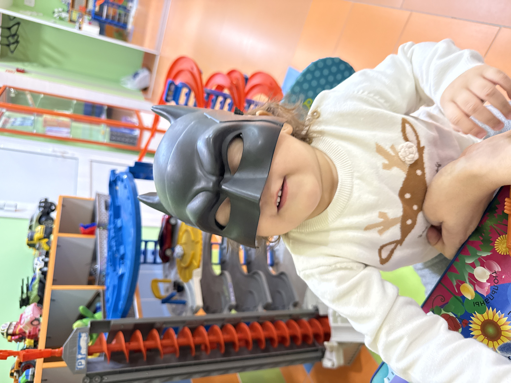 A young child, likely a toddler or preschooler, is depicted in an indoor setting from a tilted perspective. The child wears a grey plastic Batman mask over the upper half of their face, revealing a wide, joyful smile. They are dressed in a white long-sleeved sweater featuring a brown deer design on the front, adorned with a small 3D floral embellishment. An adult's hand is visible beside the child, holding a vibrant children's book. The book features colorful illustrations of sunflowers and tulips, with visible Cyrillic text including "ПОДСОЛНУХИ" (Sunflowers) and "ТЮЛЬПАНЫ" (Tulips), consistent with the location context of Tobolsk, Russia.
The background showcases a brightly colored children's play area, suggesting a daycare, kindergarten, or public play center. Various toys and play structures are visible, including orange ramps, blue plastic components, and shelves stocked with small vehicles and other play items. A prominent grey and orange toy with a spiral conveyor mechanism is in the foreground. The floor appears tiled, and parts of the walls are painted a light green. The overall scene conveys a cheerful and playful atmosphere, with the child seemingly engaged in imaginative play.
