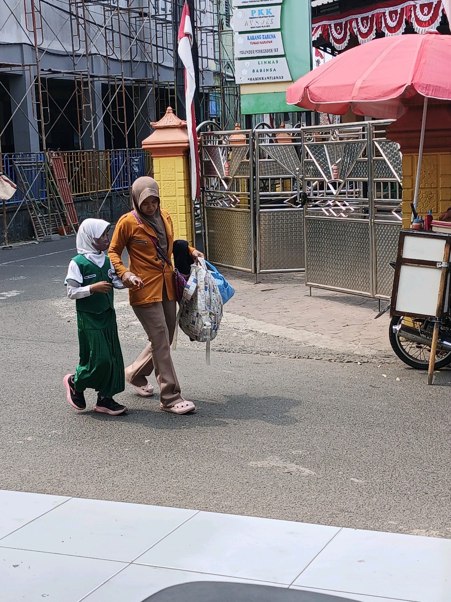A woman wearing a tan headscarf, an orange jacket, and brown pants walks on a paved road in Ketegan, Indonesia, holding a bag. Beside her, a young girl wearing a white headscarf and a green dress walks, holding the woman's hand. In the background, there are buildings under construction with scaffolding, a metal gate, and a red umbrella. The road is made of asphalt, and a sidewalk with white tiles is visible in the foreground.