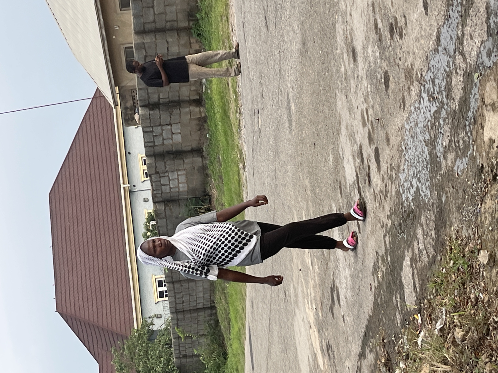 In Abuja, Nigeria, a woman wearing a grey t-shirt, black trousers, and a white and black patterned headscarf walks on a paved road. She is wearing pink and white sandals. In the background, a man in a black t-shirt and khaki trousers looks at his phone while standing near a low brick wall. Behind the wall are houses with brown and grey roofs and some green foliage. The sky is overcast.