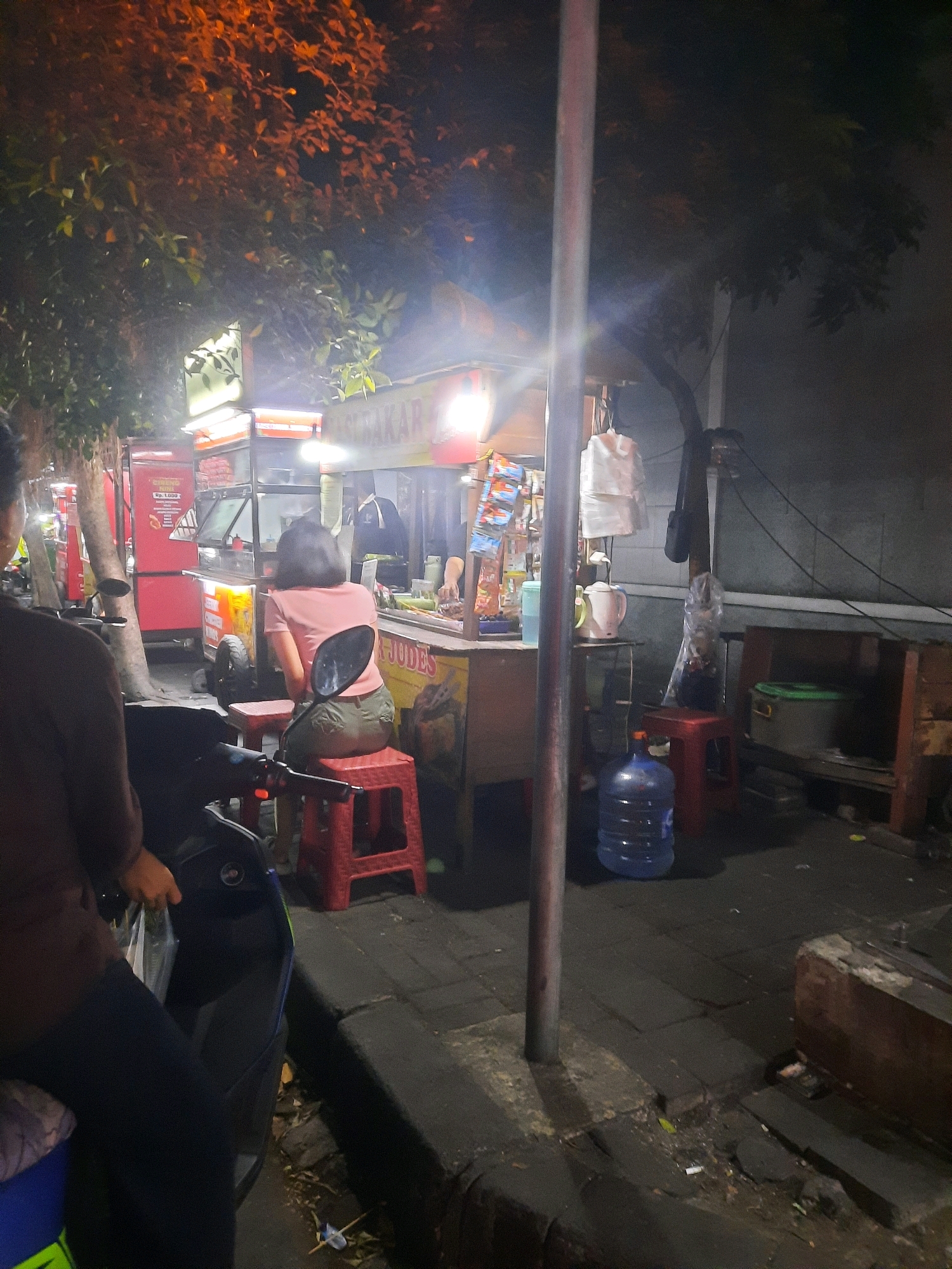 The image depicts a nighttime street scene, likely in North Jakarta, Indonesia, near a food stall. A woman with dark hair, wearing a pink t-shirt and green shorts, is seated on a red plastic stool in front of a food stall. The stall has a yellow and orange awning and appears to be selling food, with various items displayed, including packets of snacks. A large blue water jug is visible on the ground next to the stall. In the foreground, the back of a motorcycle and a person on it are visible, suggesting the woman may be waiting for or has just arrived via motorcycle. The environment is paved with brick-like tiles, and streetlights cast an orange glow, illuminating the surrounding trees and the side of a building. The overall impression is that of a typical urban street food vendor operating at night.