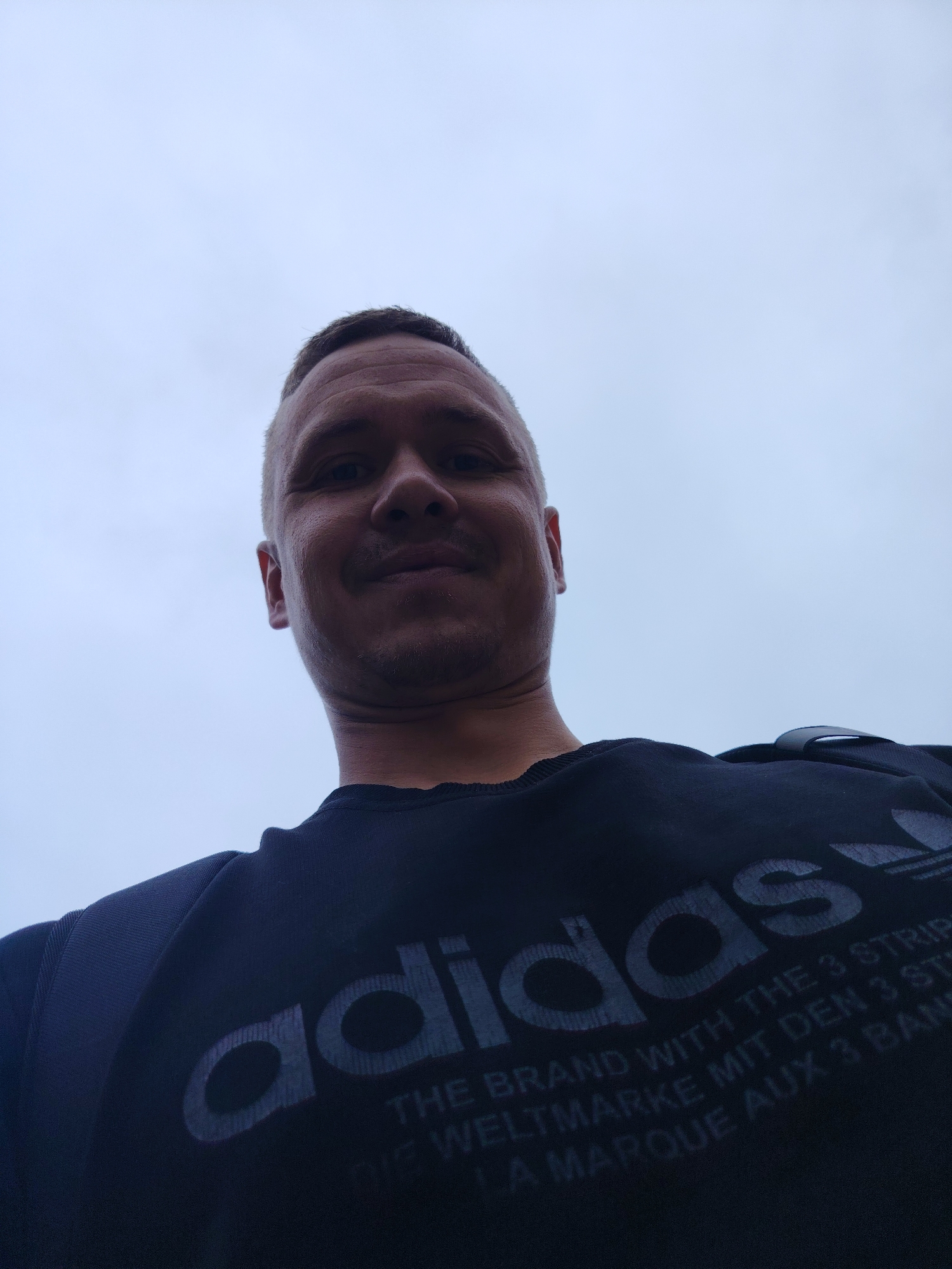A low-angle, outdoor, waist-up, medium shot captures a man with short hair and stubble, wearing a dark sweatshirt with the Adidas logo and text. The man is looking up towards the camera with a slight smile. The background is a light gray, overcast sky. The image appears to have been taken in the city of Chaikovsky, Russia, possibly on a street or public area, given the casual attire and open sky.