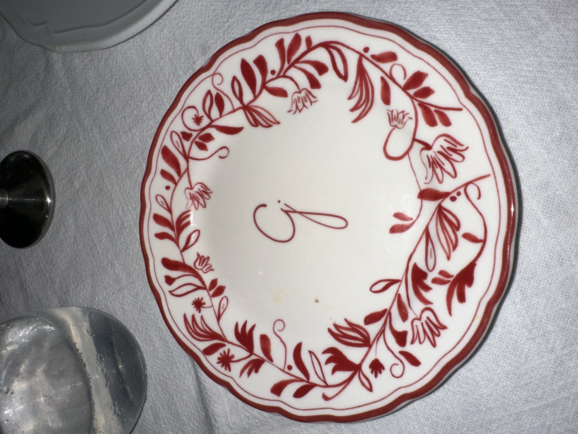 The primary subject of the image is a white ceramic plate featuring a decorative red design. The plate has a scalloped edge and is adorned with a continuous wreath-like pattern of red foliate and floral motifs. These motifs include stylized leaves, small bell-shaped flowers, and tiny star-like blossoms, all rendered in a deep red hue against the bright white background of the plate.
In the center of the plate, a prominent red calligraphic initial "J" is visible, suggesting a personalized item. The plate rests on a light-colored, textured fabric surface, possibly a tablecloth, which appears to be white or a very light grey.
In the blurred background on the left side, several other objects are partially visible. These include a portion of another smooth, white ceramic dish or bowl, a dark metallic cylindrical object (perhaps a utensil handle or candle holder), and a translucent, textured, rounded object, possibly a glass or decorative item.
No human subjects are present, and no activity or event is overtly taking place; the scene appears to be a still life. The lighting is soft and even, suggesting an indoor setting. There are no direct visual cues within the image itself to indicate the provided location context of North Kuta, Indonesia. The visible text is the single calligraphic letter "J" on the plate.