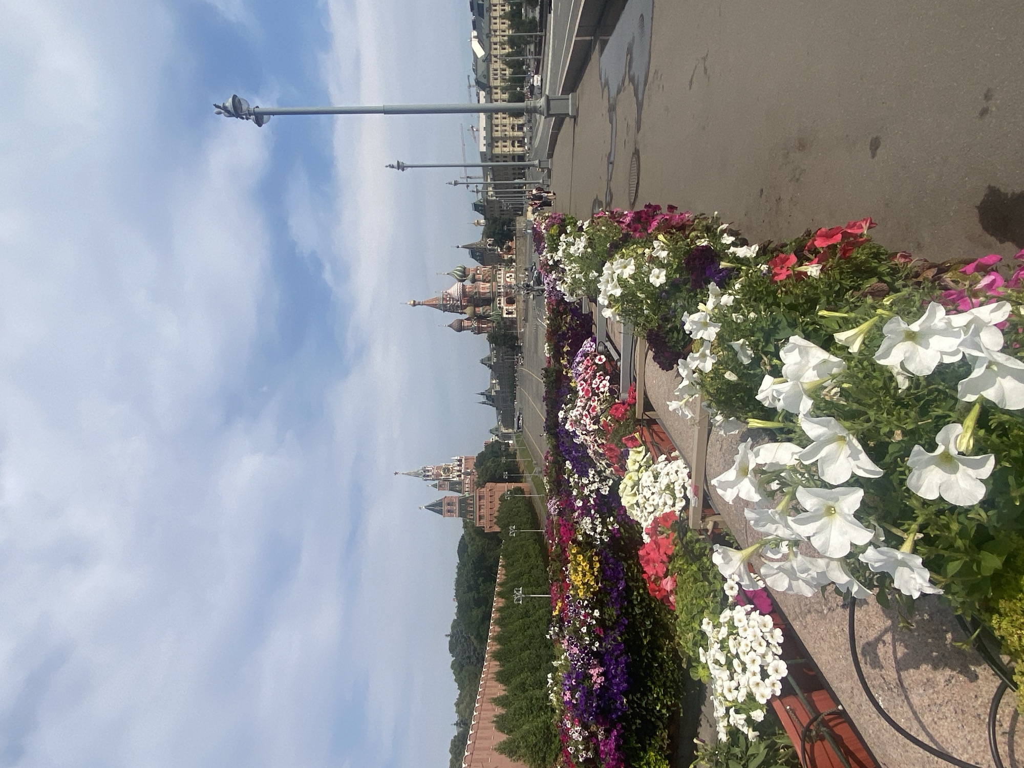 This is a photograph of a flower bed along a walkway in Moscow, Russia, with St. Basil's Cathedral and the Kremlin visible in the background. The foreground is filled with numerous blooming flowers in various colors, including white, pink, purple, and red petunias. A stone embankment borders the flower bed. To the right of the flowers is a paved walkway, and beyond that, a road with vehicles. Several lampposts line the walkway. In the distance, the iconic domes of St. Basil's Cathedral and the towers of the Kremlin are prominent. The sky is partly cloudy, with a bright blue sky and white clouds. The scene appears to be set on a sunny day.