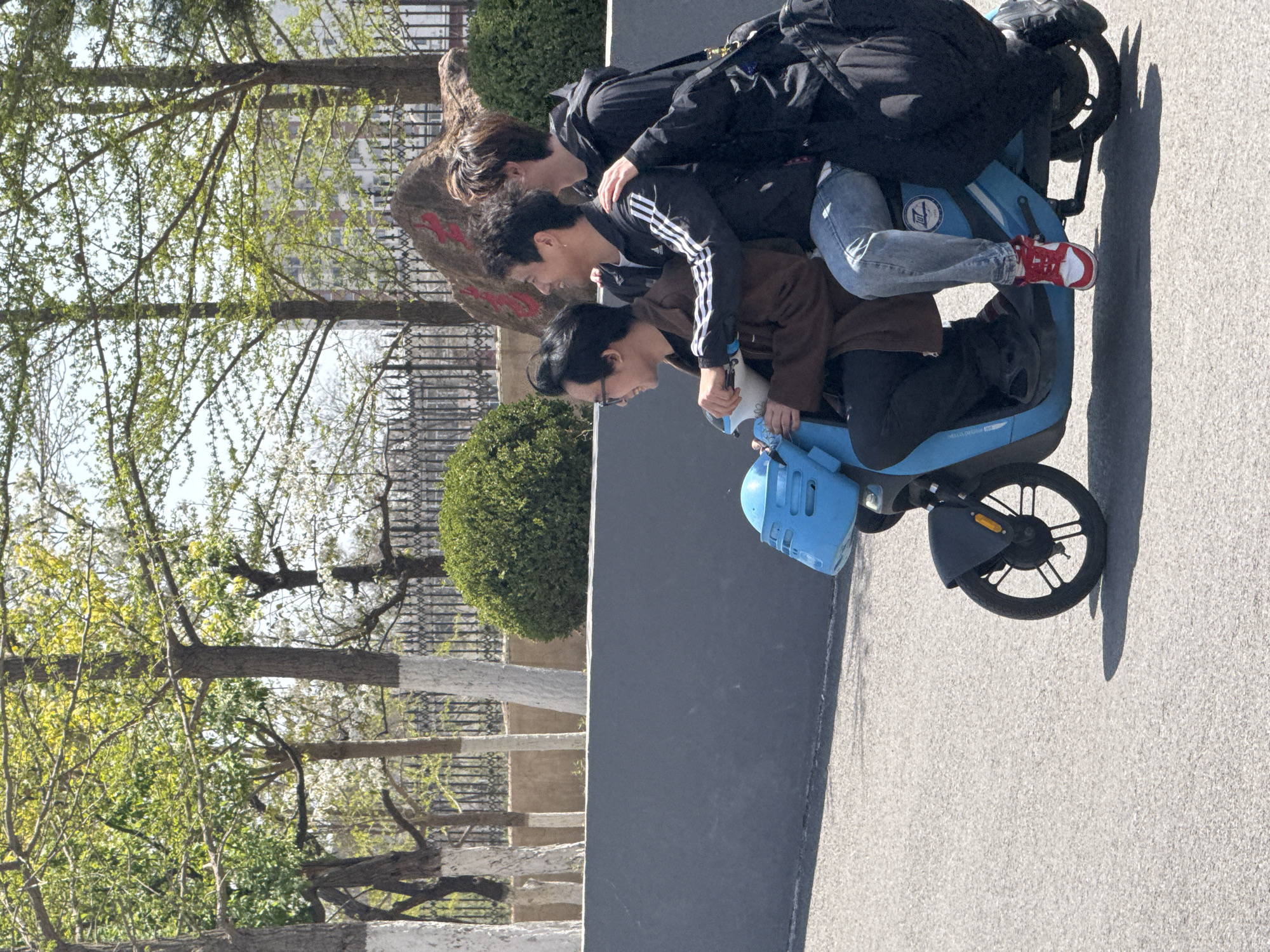 Three people are riding a blue electric scooter on a paved surface. The person in front is wearing glasses and a brown jacket. The person in the middle is wearing an Adidas jacket with white stripes. The person in the back is wearing a black jacket and jeans. The scooter has red and white sneakers on its platform. Behind the scooter is a low concrete wall with green bushes. Trees are in the background. The photo was taken in Zhongtun, China.