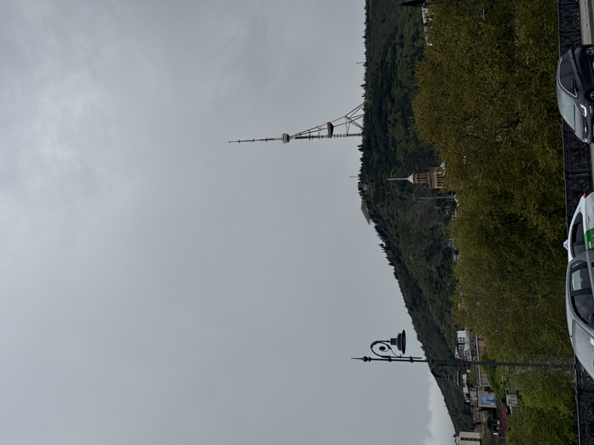The image presents a view of the city of Tbilisi, Georgia, taken on an overcast day. In the background, a tall television tower stands atop a hill densely covered in green foliage. Closer to the viewer, a street with parked cars is visible. A vintage-style street lamp is also visible on the side of the road.