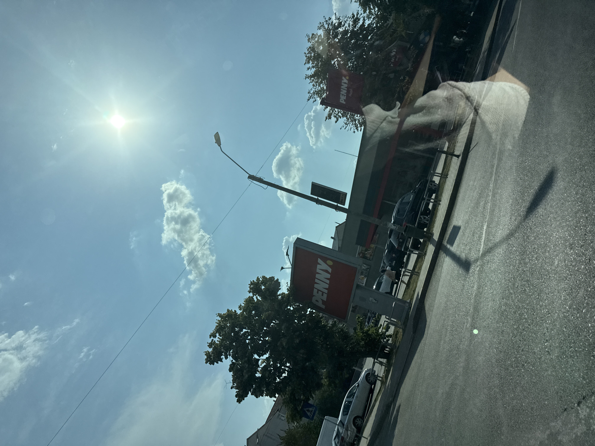 This is a medium shot, taken from a vehicle, of a street scene in Huși, Romania. The sky is bright blue with scattered white clouds and the sun is shining intensely. A street light with a curved arm extends from the right side of the frame towards the center. In the middle ground, there is a Penny Market store with a red sign. A large tree with dense green foliage stands to the left of the Penny Market sign, partially obscuring it. Cars are parked on the side of the road in front of the store. In the foreground, the asphalt road is visible, with a slight greenish reflection in the middle. There is a reflection of a light-colored fabric, possibly clothing, on the surface from which the photo is taken.