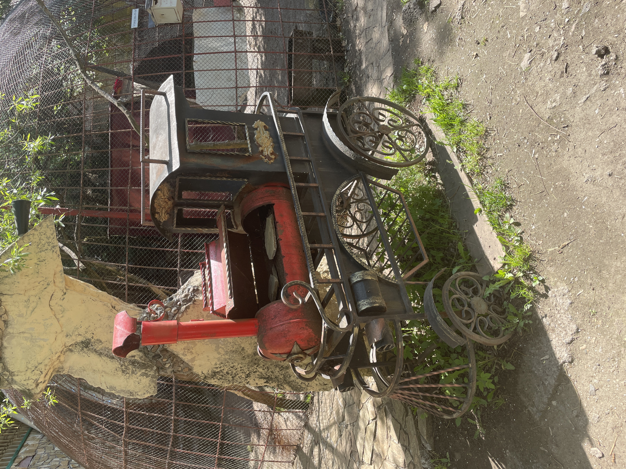 The image shows a decorative, rusted metal train engine placed near a fence and landscaping in what appears to be an outdoor area. The train is painted in a combination of red and dark metal colors, with ornamental wheels and details. The ground is a mix of dirt and small stones with patches of green vegetation. Based on the image context, it is likely that this is an example of urban art in Perm, Russia.