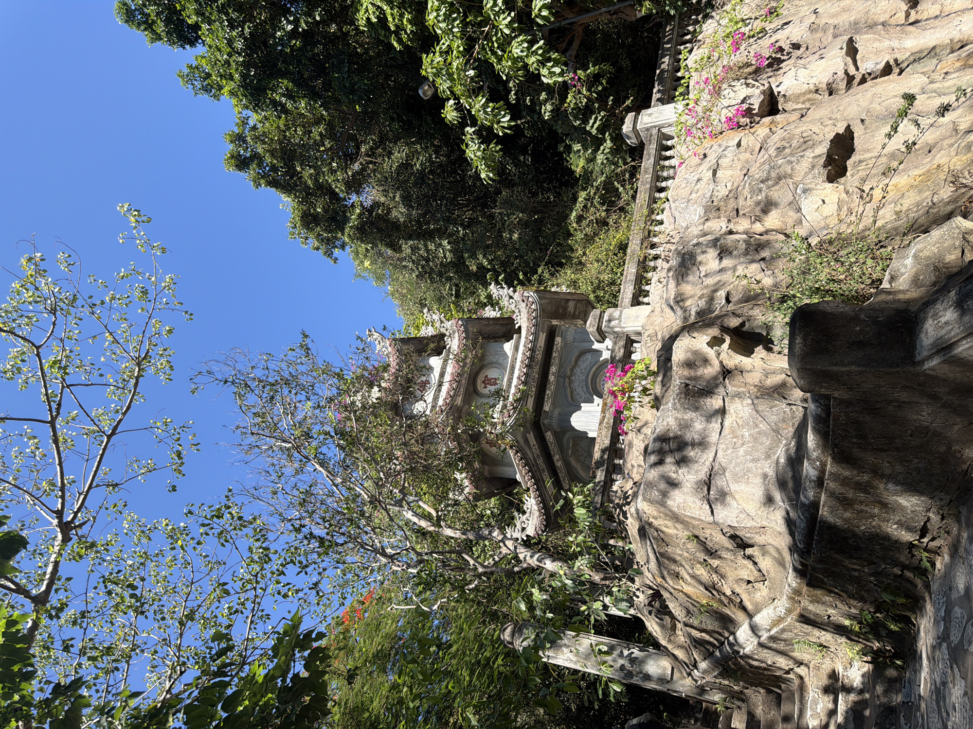 An outdoor scene captures a multi-tiered, ornate pagoda or temple structure nestled into a rugged, rocky cliffside, surrounded by lush vegetation under a clear blue sky. The architectural structure, likely a shrine, is made of light-colored stone and features intricate decorative elements, including circular medallions and elaborate sculptural details. Patches of vibrant pink, bougainvillea-like flowers cling to the rocky face and parts of the building, adding splashes of color to the predominantly stone and green palette.
Dense green foliage and various trees frame the structure, with some tree branches and leaves silhouetted against the bright, cloudless blue sky, which occupies the upper portion of the image. The natural rock formations are prominent, displaying textured surfaces and varying shades of gray and brown. Stone railings or balustrades are visible, suggesting pathways or stairs integrated into the landscape.
The bright sunlight casts distinct shadows across the rocks and the structure, indicating a clear, sunny day. No people are visible, contributing to a tranquil and serene atmosphere. The scene depicts a harmonious blend of natural beauty and traditional architecture, characteristic of a sacred or historical site found in a mountainous or hilly region, consistent with the provided location context of Đà Nẵng, Vietnam. No visible text is present in the image.