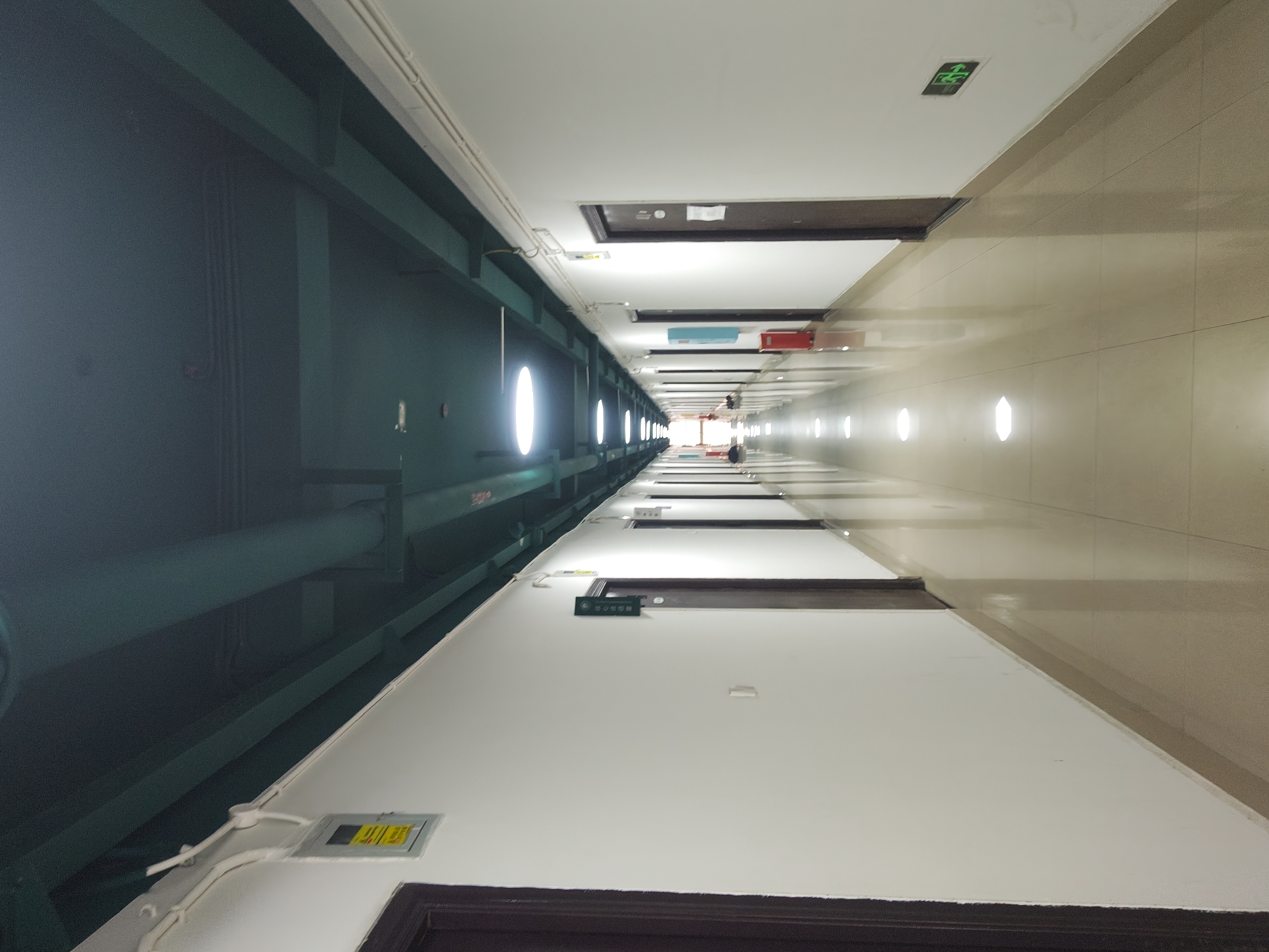A long, brightly lit hallway stretches into the distance, lined with closed doors on both sides. The walls are painted white, and the floor is a polished, reflective tile that mirrors the circular ceiling lights. The ceiling itself is dark and appears to house exposed pipes and conduits, painted in a dark green hue. On the left wall, a yellow and black electrical panel is mounted, and further down, a dark brown door with a nameplate is visible. On the right wall, a series of similar dark brown doors are present, one with a small white sign and another with a green exit sign. In the distance, figures can be seen walking down the hallway. The overall impression is one of a modern, functional building corridor, likely in a commercial or institutional setting. This scene is located in the Chongqing High-tech Zone, China.