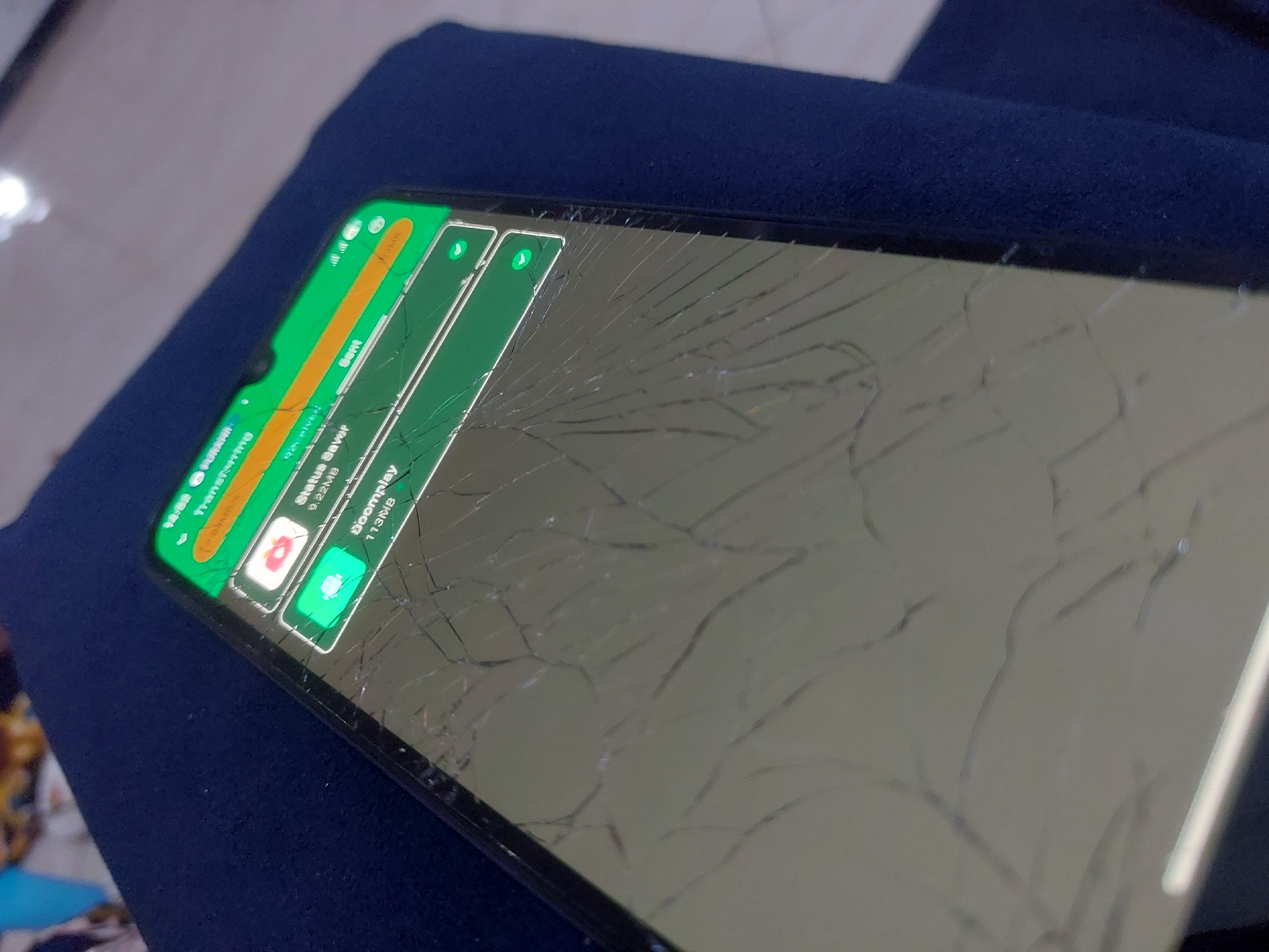The image shows a smartphone with a heavily cracked screen lying on a dark blue cloth or surface. The phone is displaying notifications, possibly related to file transfers or downloads. The screen damage obscures much of the underlying display. No people are visible. There is no information available to indicate the location is Dar es-Salaam, Tanzania.