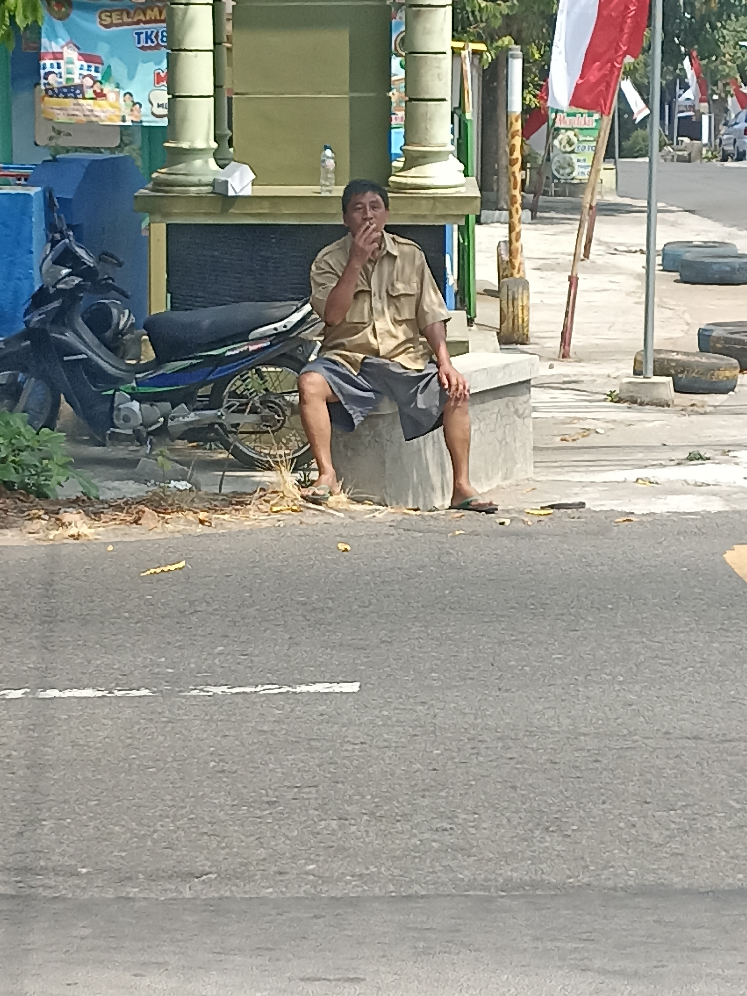 A man in a tan shirt and dark shorts sits on a concrete ledge, smoking a cigarette. He is positioned next to a motorcycle. In the background, a banner displays Indonesian text, and a flagpole with an Indonesian flag is visible. The scene appears to be in Pulisen, Indonesia, given the presence of Indonesian cultural elements and the context provided. The environment suggests an outdoor, possibly urban setting, with a road and roadside elements like discarded tires.
