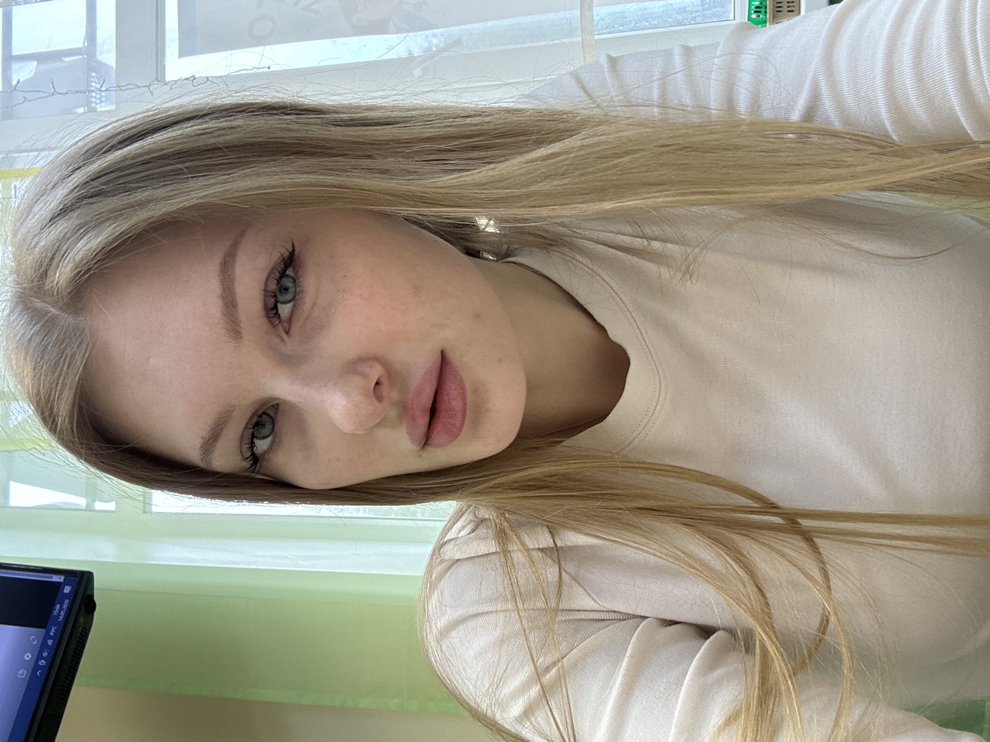 The image is a close-up, selfie-style shot of a young woman with long, blonde hair and light eyes. She is wearing a light-colored, long-sleeved shirt. The background is blurry, but what is visible suggests an indoor setting, possibly an office or study. A window with light-colored curtains is visible, along with a section of a computer screen. The computer screen has visible Russian text at the bottom. Based on the text visible, the location could be Perm, Russia.