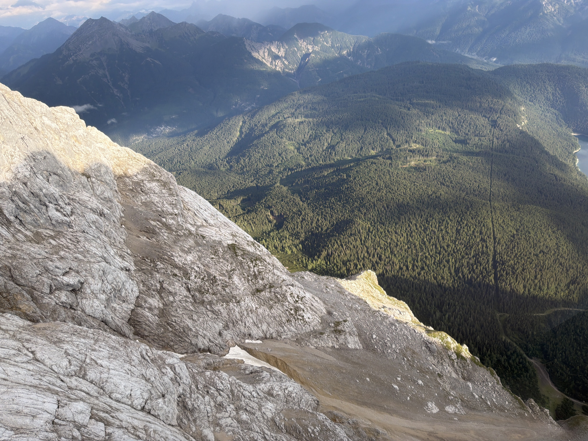 This is an aerial view of the Zugspitze mountain in Grainau, Germany, taken from a high vantage point looking down. The foreground is dominated by the rugged, grey, rocky terrain of the mountain slope, with a sharp drop-off visible. The slope is partially illuminated by sunlight, creating distinct shadows. In the midground and background, rolling green hills covered in dense coniferous forest stretch out towards distant, hazy mountain ranges. Sunlight breaks through the clouds in patches, casting dramatic shadows across the landscape. A narrow body of water, likely a lake, is visible in the far right background. There are no people or animals in the image.