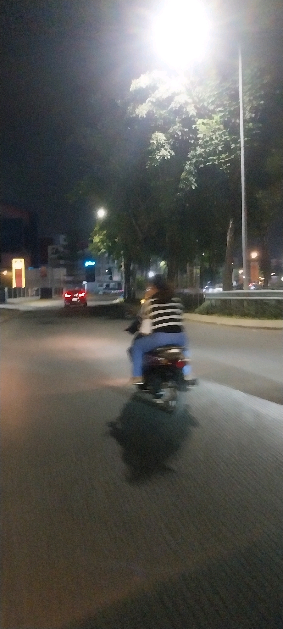 The image depicts a street scene at night in Medan, Indonesia, characterized by motion blur suggesting movement. A person is riding a motorcycle, viewed from behind. The rider is wearing a horizontally striped shirt and blue pants. The motorcycle itself is dark-colored. The background shows trees and streetlights, with a red car visible in the distance on the road. Buildings are present on the left side of the street, with some illuminated signs. The overall lighting is low, with a bright street lamp at the top of the frame casting a glare. The road surface is visible, and a shadow of the motorcycle and rider is cast on the ground.