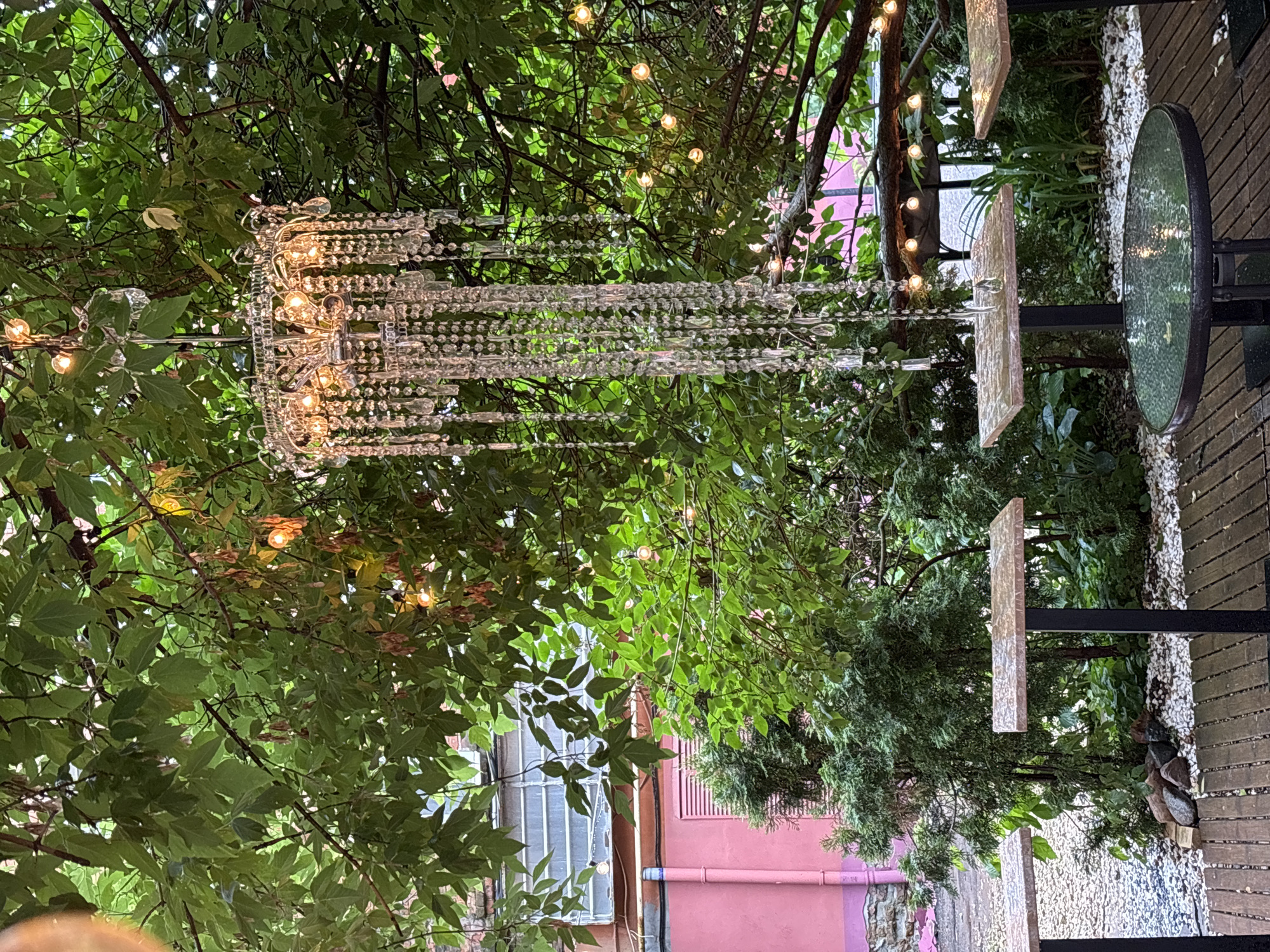 The image depicts an outdoor seating area, likely a cafe or restaurant, adorned with a decorative crystal chandelier hanging from tree branches. The area is furnished with several small tables, including a round glass-topped table in the foreground and two square-topped tables with dark metal legs. The ground is covered with wooden planks and white pebbles, with some rocks scattered near the tables. Lush green foliage from trees and bushes surrounds the seating area, creating a natural, shaded ambiance. String lights are also visible amongst the branches, suggesting an evening setting. In the background, a portion of a building with pink and grey walls and a window is visible. The scene is located in the city of Nizhny Novgorod, Russia. No people are present in the image.