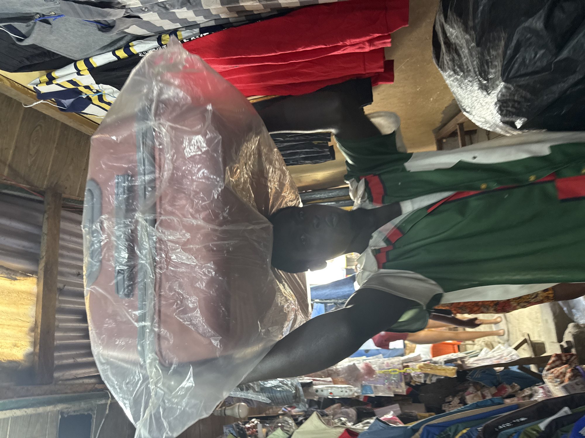 A young Black male is captured from the chest up, walking in a market stall, likely in Lekki, Nigeria. He is carrying a large, red suitcase, covered in clear plastic, balanced on his head. The man is wearing a green and white shirt with red trim, and his head is tilted back slightly.
The environment appears to be an outdoor market with a corrugated metal roof supported by wooden beams. Clothing is hanging on racks and shelves to the right of the man, including red shirts and striped garments. To his left, various items are displayed on shelves, and the background shows more of the market's interior with hanging goods. A light bulb hangs from the ceiling, casting a warm glow. The ground is not clearly visible.