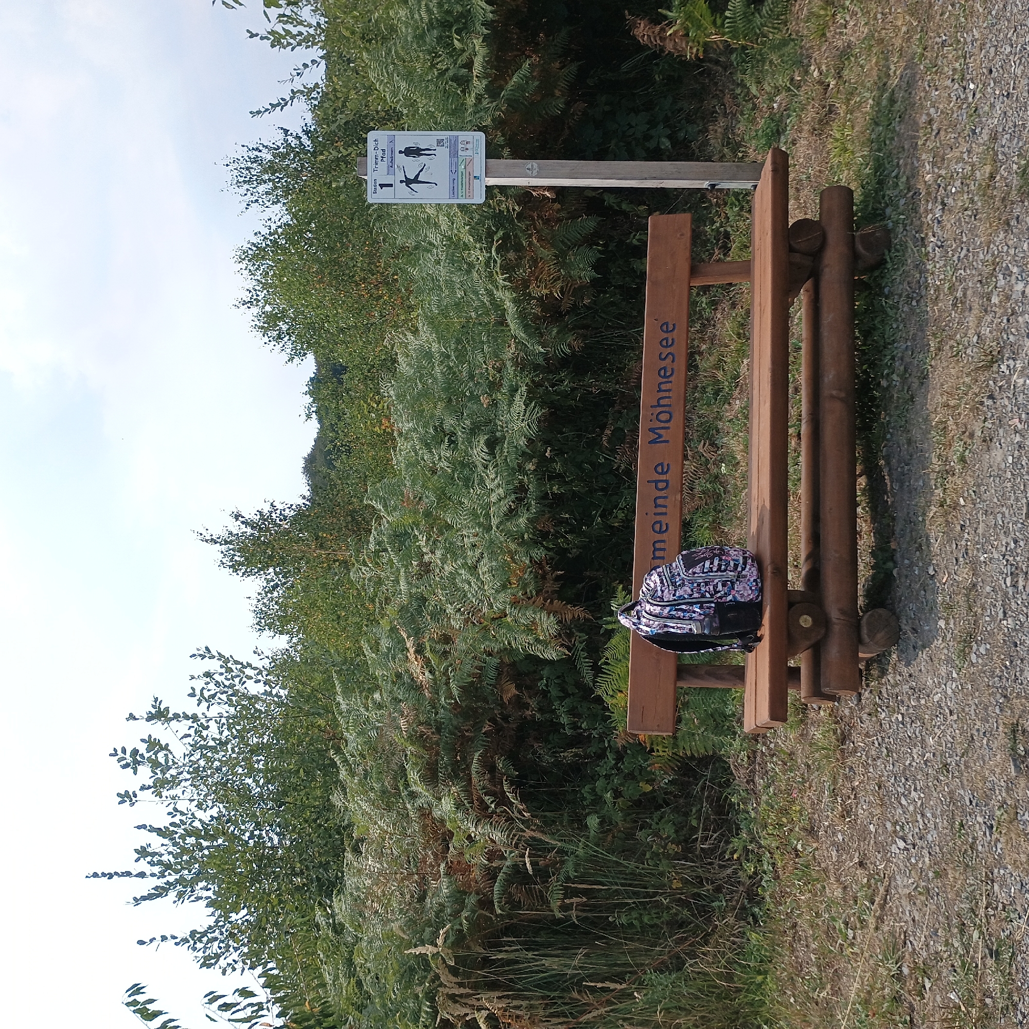 A wooden bench with the text "Gemeinde Möhnesee" carved into it sits on a gravel path in a natural environment. A backpack with a colorful pattern rests on the left side of the bench. To the right of the bench is a wooden post with a sign that reads "Station 1 Trimm-Dich-Pfad". The sign features illustrations of people exercising. The background consists of dense green foliage, including trees and ferns, under a partly cloudy sky. The scene appears to be located in Möhnesee, Germany, given the inscription on the bench.