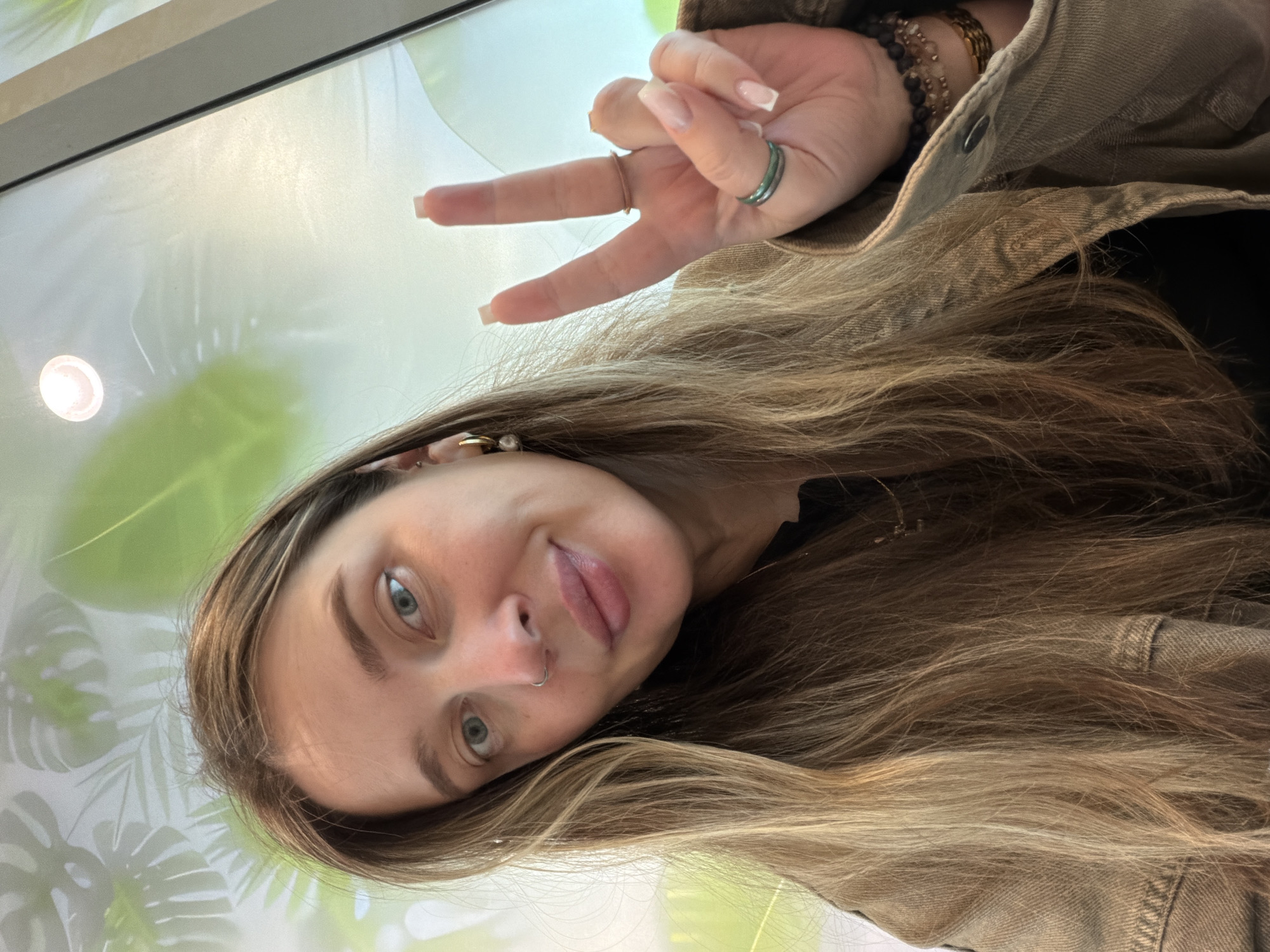 A woman with long brown hair poses indoors, sticking out her tongue slightly and holding up a hand in a peace sign. She wears a denim jacket and multiple rings. She has a nose piercing. The background appears to be a white and green surface, possibly a wall or large table. The location in Domodedovo, Russia cannot be determined based on the image.