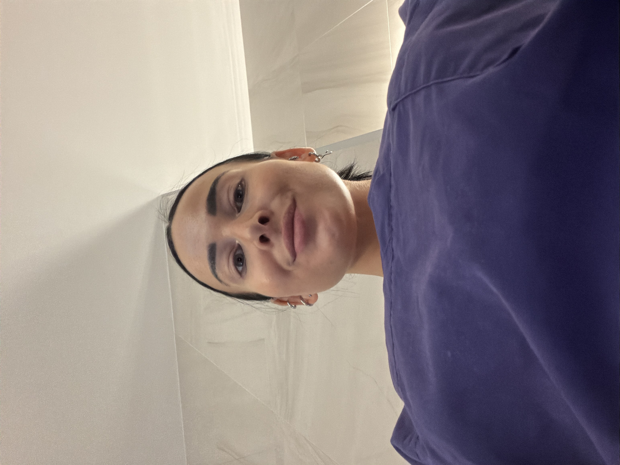A low-angle, close-up shot captures a woman's face from below as she looks up at the camera. She has dark hair pulled back, styled in a ponytail, and is wearing subtle makeup. Multiple silver hoop earrings are visible on her left ear. She is wearing a purple scrub top. The background consists of a light-colored tiled wall and a textured ceiling. The lighting suggests an indoor environment. There is no information in the image to suggest the location is Saint Petersburg, Russia.