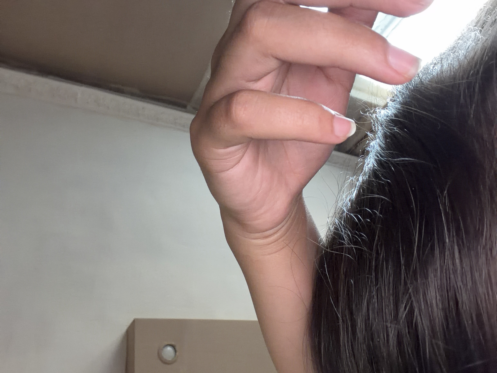 A close-up shot shows a person's left arm and hand resting on their dark hair. The arm is bent at the elbow, with the hand resting on top of the head, fingers curled slightly. The skin on the arm and hand is lightly tanned and shows natural creases. The fingernails are short and clean. The hair is dark brown and appears to be straight and slightly damp, with some finer strands visible. In the background, a light-colored wall with a faint horizontal line is visible, along with a corner of a light brown piece of furniture with a round, circular object on it, possibly a light fixture or decorative element. A vertical edge of a window or door frame separates the wall from a darker background. The scene is lit by a light source coming from the right, casting some shadows on the arm and hand. The location is identified as Razhevo Konare, Bulgaria.