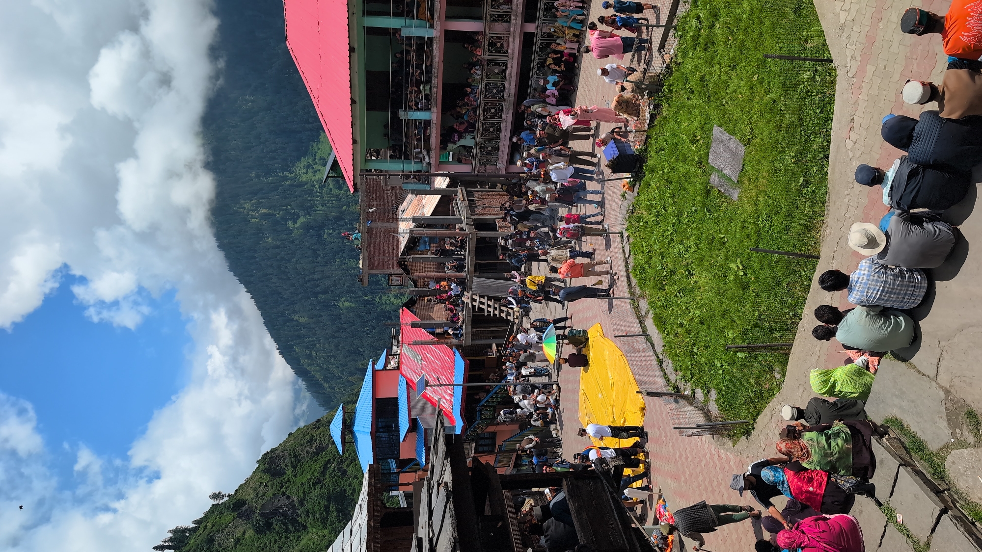 The image depicts a vibrant community gathering in the village of Malana, India, likely during a local festival or event. The scene is set in an open courtyard surrounded by traditional multi-story buildings with colorful roofs, predominantly red and blue. The architecture suggests a mountainous region, with lush green hills and pine forests visible in the background, under a partly cloudy sky.
In the foreground, a group of people are seated on stone steps, facing away from the camera, watching the activities in the courtyard. They are dressed in a mix of traditional and modern attire.
The courtyard itself is bustling with activity. A large, bright yellow tarp is spread out on the ground, with several individuals standing or sitting around it. Many other people are milling about, some in small groups engaged in conversation, while others appear to be observing the event. There are also some children present, adding to the lively atmosphere.
Several buildings in the background feature balconies where more people are gathered, suggesting a communal viewing area or part of the celebration. The overall impression is one of a well-attended, traditional gathering in a scenic, rural setting.