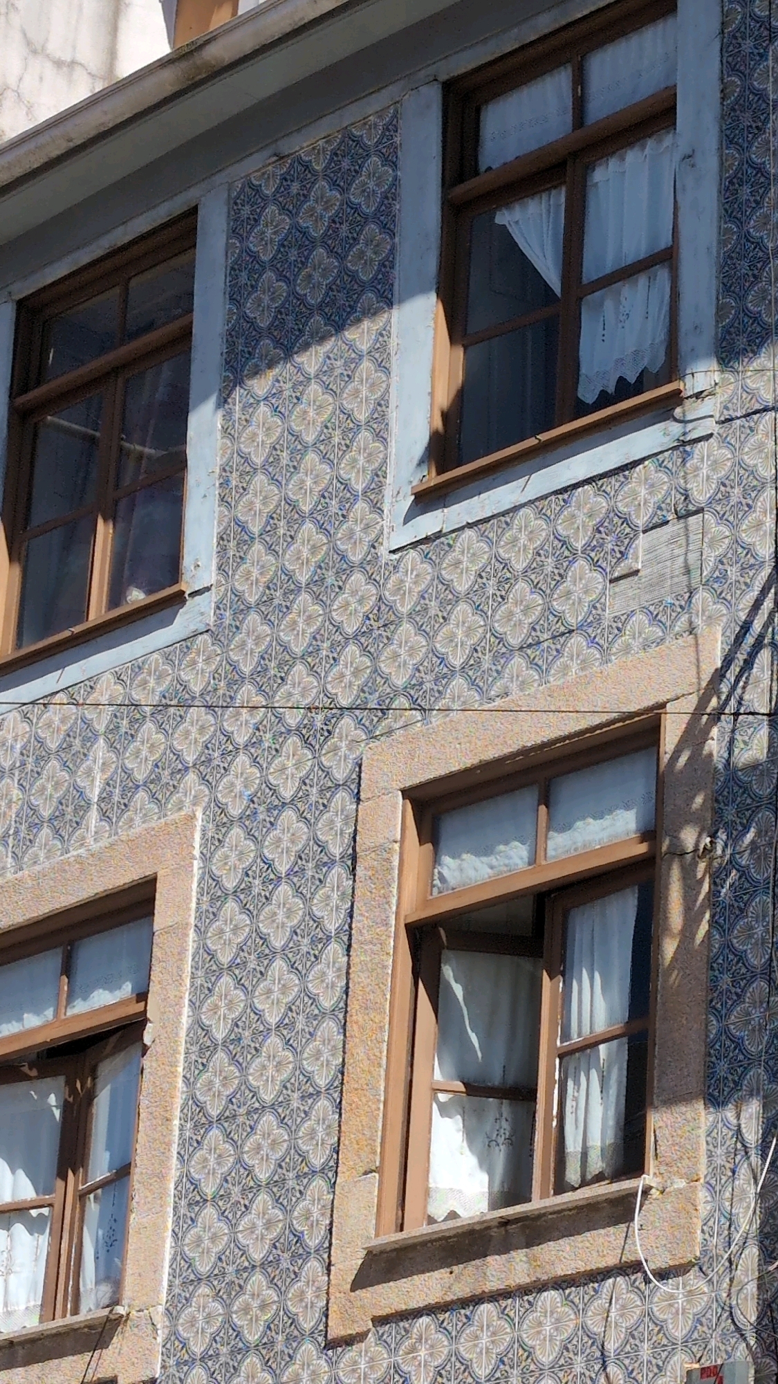 This is a close-up, eye-level exterior shot of a building's facade, likely in Porto, Portugal, given the architectural style and the common use of azulejo tiles. The building features multiple windows with wooden frames and white lace curtains. The walls are adorned with intricate blue and white azulejo tiles, arranged in a repeating pattern of quatrefoil shapes. The tiles are partially shaded by the sun, indicating it is daytime.
There are no people visible in the image. The activity depicted is simply the static presence of the building itself, showcasing its architectural details. Notable details include the weathered appearance of the building, with some areas of the tiled surface showing wear and tear. The windows are set into frames made of what appears to be stone or a similar textured material. The sunlight casts strong shadows on the tiled walls, suggesting a sunny day. The overall impression is one of an older, traditional European building. No text is visible in the image.