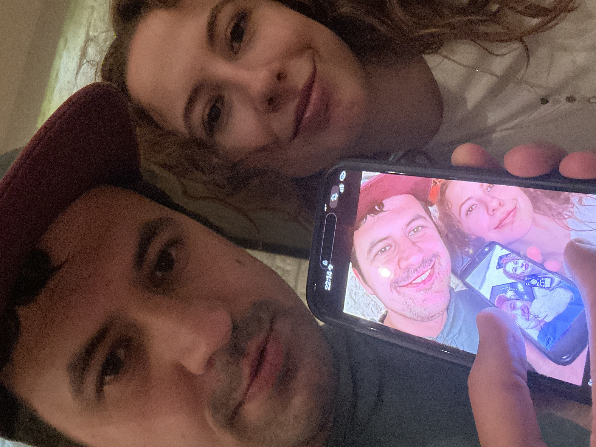 An intimate selfie captures a man and a woman lying down indoors. The man, wearing a dark red cap, holds a smartphone in his right hand, angled to include himself and the woman behind him in the current photo. Both the man and the woman are smiling warmly, conveying a sense of happiness or contentment.
The smartphone screen displays another image of the same two individuals, also smiling, creating a recursive "photo within a photo" effect. The image on the phone screen shows a similar composition, suggesting it's another selfie they previously took, and within that displayed image, another phone with an even smaller image of them can be faintly seen, adding another layer to the recursion. The phone screen clearly shows the time "22:10".
Unexpectedly, three light-brown eggs are visible resting on a surface directly behind the top edge of the smartphone. The setting appears to be a cozy indoor environment, possibly a bedroom, given the relaxed poses and what might be bedding or cushions in the background. The scene is set in Podgorica, Montenegro.