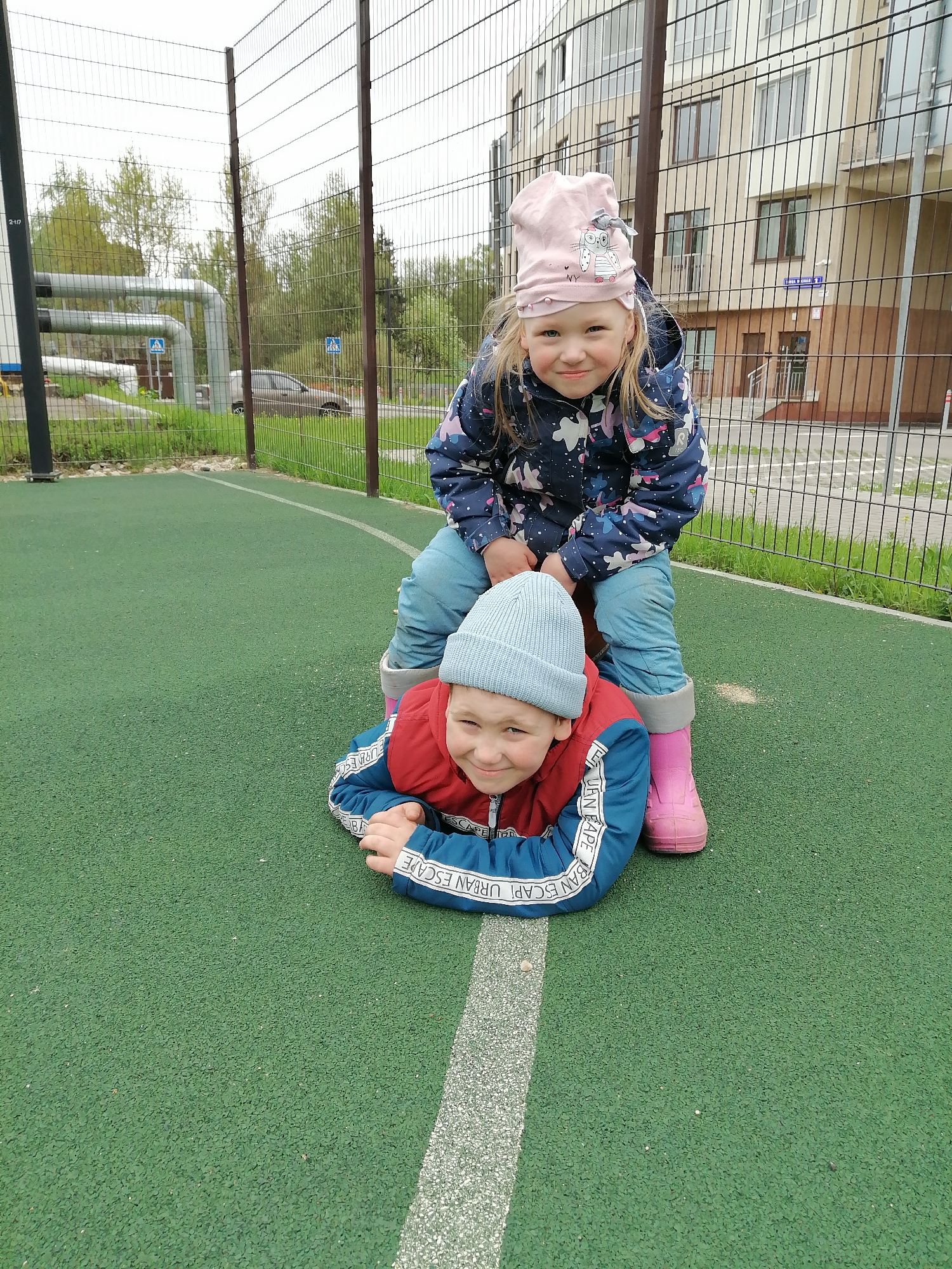 Two children are playing on a green surface with a white line marking, potentially a sports field. A girl with blond hair, wearing a pink hat and a blue jacket with a starry pattern, is sitting on the back of a boy who is lying face down on the green surface. The boy is wearing a blue hat, a red and blue jacket with the words "URBAN ESCAPE" on the sleeves. The girl is wearing pink boots. A metal fence is visible in the background, along with trees and a building. A street sign is visible on the building indicating the location is in Trubacheyevka, Russia. A pipe and other building are also visible in the background.