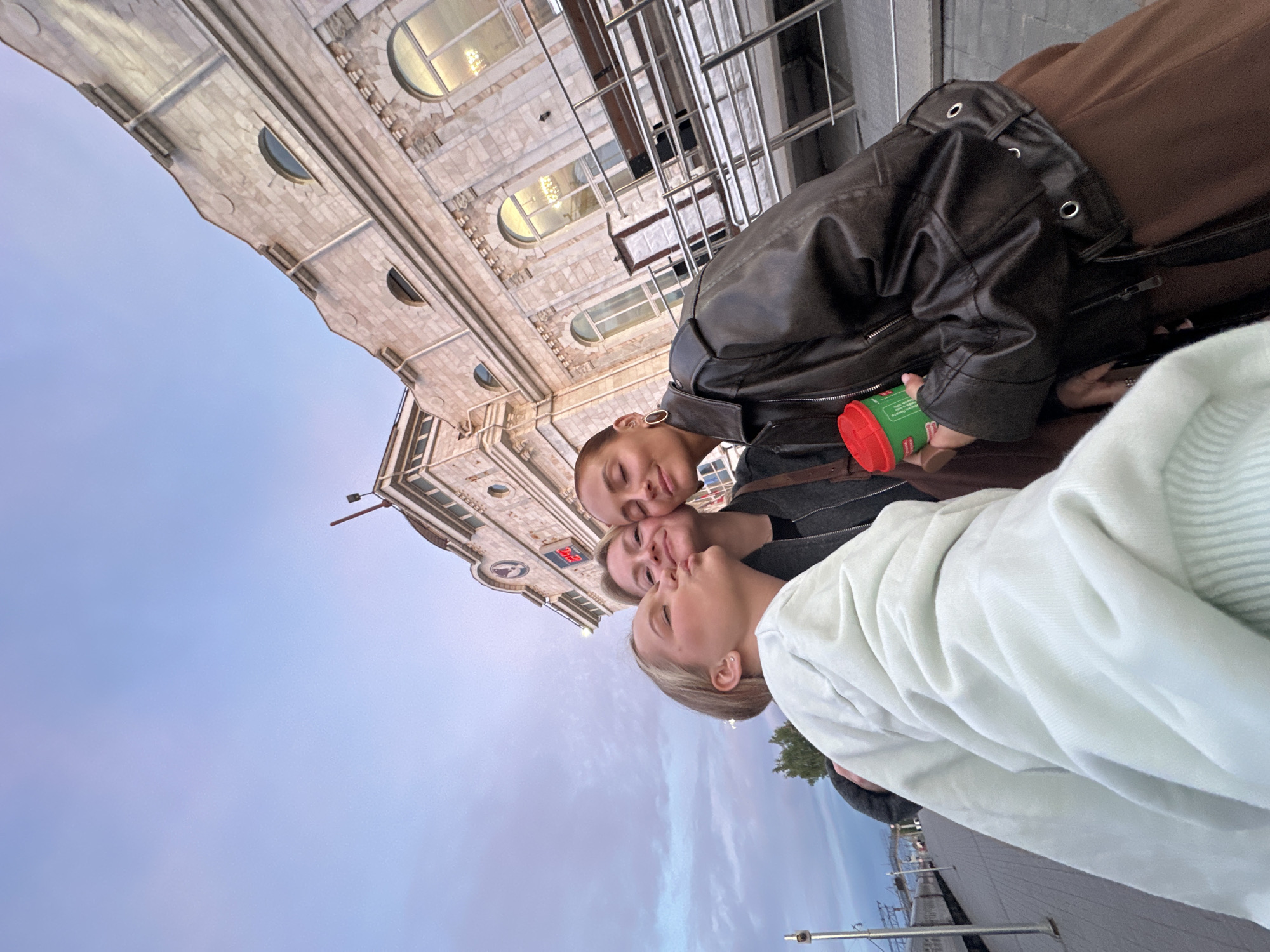 Three people are taking a selfie in front of a building with a clock on it in Izhevsk, Russia. The building is light colored with arched windows and decorative stonework. The sky is light blue with some clouds. The person in the front is wearing a white sweatshirt. The person in the middle is wearing a dark gray jacket. The person in the back is wearing a black leather jacket and a brown shirt or jacket. They are holding a red and green container. The location is likely near the Izhevsk railway station, based on the architectural style of the building in the background.