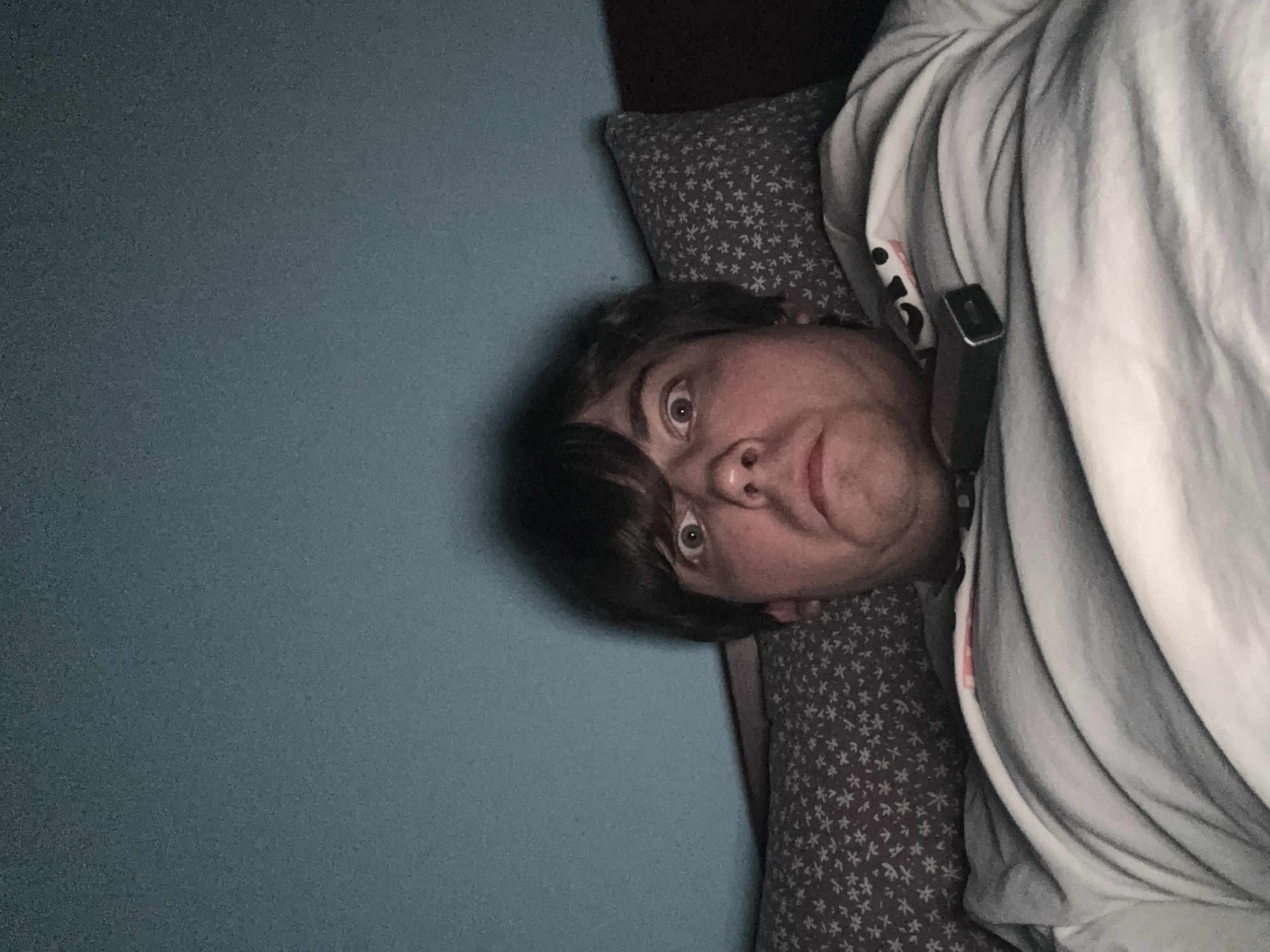 A person is lying in bed, facing the camera. The person has light brown hair and is looking up with wide eyes, a surprised expression. They are wearing a grey and white striped t-shirt. A black object, possibly a vape device, is resting on their shirt. The background consists of a teal wall and two pillows, one with a white floral pattern on a grey background, and the other a darker color. The lighting is dim, creating a shadow behind the person's head. The image appears to have been taken in a bedroom.