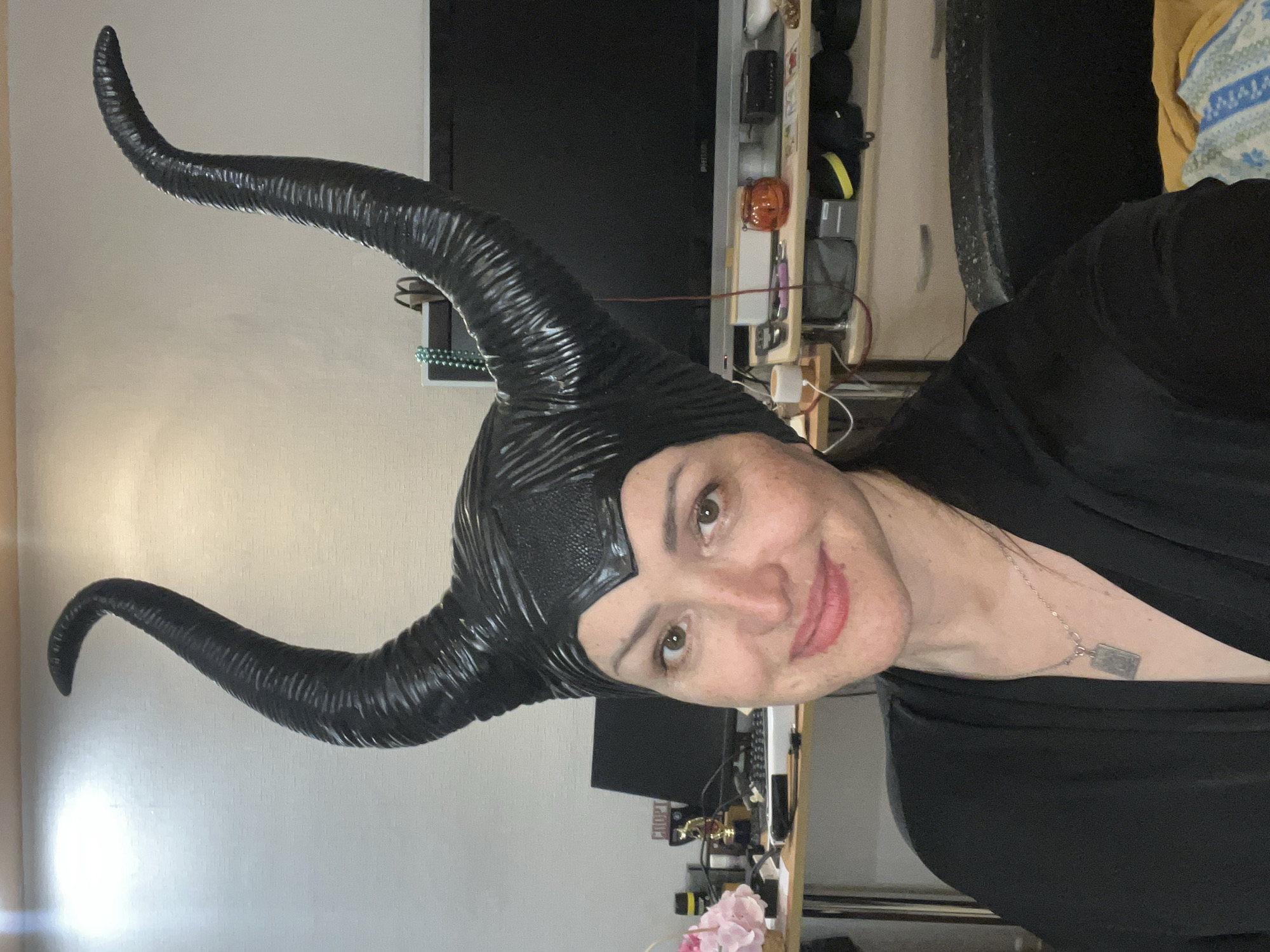 A woman is posing in front of a desk, wearing a black Maleficent horned headpiece. She is wearing a black robe or garment. She is wearing a silver necklace with a square pendant. The background shows a desk with various items on it, including a keyboard, computer monitor, and decorative flowers. There is a television mounted on the wall behind the desk. The location appears to be indoors, possibly in a home or office setting. There is no indication of the city of Perm, Russia, or any location information present in the image.
