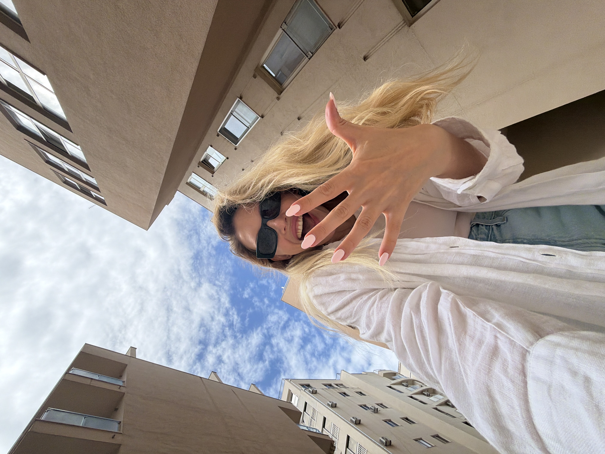A low-angle medium shot depicts a woman with long blonde hair and sunglasses extending her hand towards the camera. She wears a white linen shirt and what appears to be denim shorts. Her nails are painted a pale pink. She smiles slightly. The background features a cityscape of Budva, Montenegro, with various apartment buildings of similar beige color. The sky is partially visible with clouds.