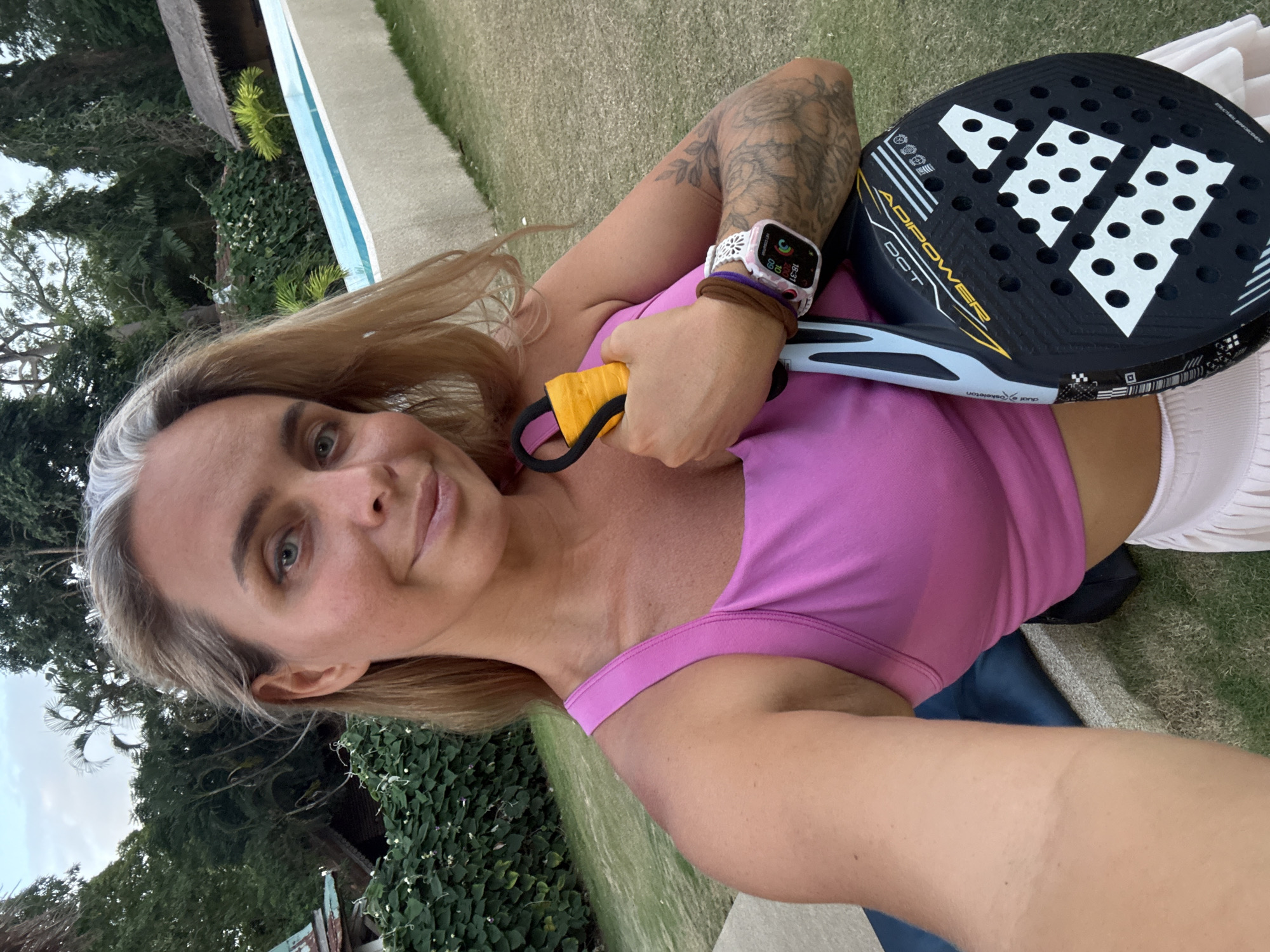 In a self-taken photograph, a woman with blonde hair and grey streaks stands outdoors in a grassy area with lush green foliage and trees in the background. To her right, a swimming pool with blue water is visible, bordered by a light-colored wall. The woman is wearing a pink tank top and a light-colored skirt. She is holding a black and yellow paddle, identified by the "ADIPOWER" logo and Adidas branding, suggesting it is a padel racket. On her left wrist, she wears a smartwatch with a white floral-patterned band. The scene appears to be in a tropical setting, consistent with the location of Ko Samui District, Thailand, possibly a resort or private residence.