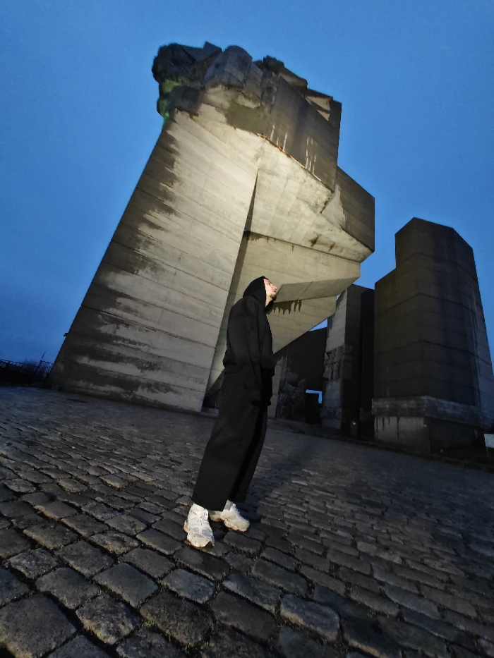 An individual stands on a cobblestone plaza at the base of the Monument to 1300 Years of Bulgaria, situated on the Shumen Plateau overlooking Shumen, Bulgaria. The scene is illuminated by low light, suggesting dusk or night, under a dark blue sky. The subject, a person wearing a black hooded sweatshirt with the hood up, black trousers, and white athletic shoes, is positioned slightly right of the frame's center. They face right and upward, with their head tilted back, gazing at the colossal concrete monument. The monument consists of multiple angular, raw concrete blocks, exhibiting visible weathering and streaks. A hint of green light or material is discernible on the uppermost concrete mass. To the right and behind the central structure, another large, block-shaped concrete element of the monument is visible. The foreground features an uneven surface paved with dark, irregularly shaped cobblestones. The image is captured from a low, upward-angled perspective with a slight rightward tilt.