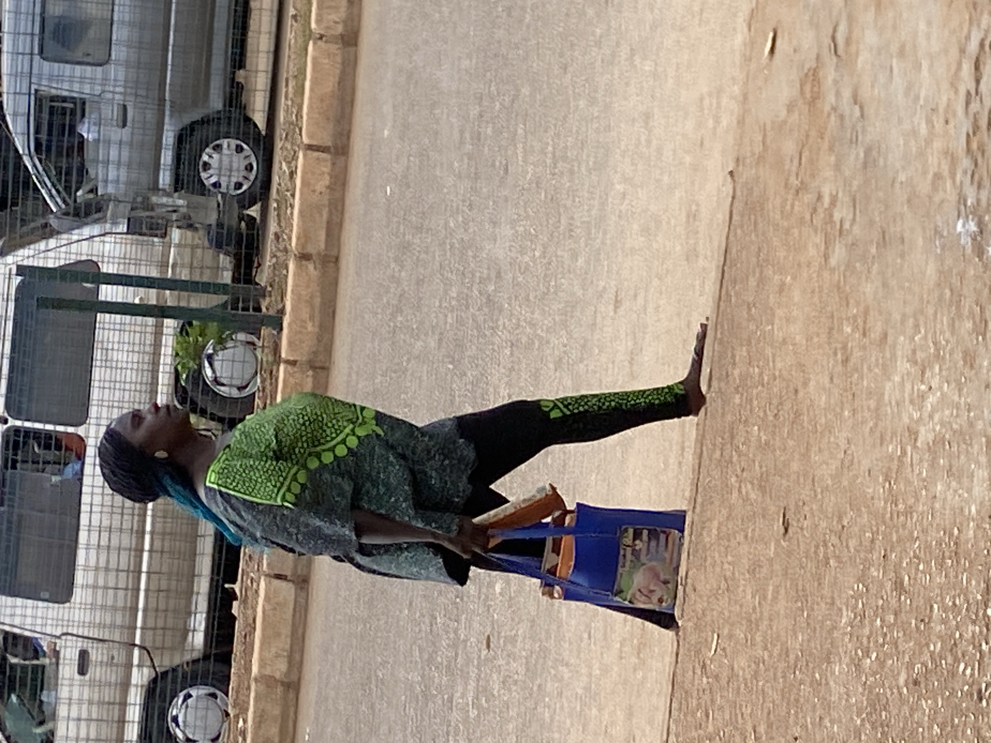 A woman is walking on a paved surface, possibly a road or sidewalk, in Bukuru, Nigeria. She is wearing a dark grey and green patterned top and black leggings with a green pattern. She is carrying a blue tote bag in her left hand, which contains some items. Her right hand is holding an orange object. The background shows a light-colored van or truck with a protective cage on its side. The ground is a mix of dirt and asphalt.