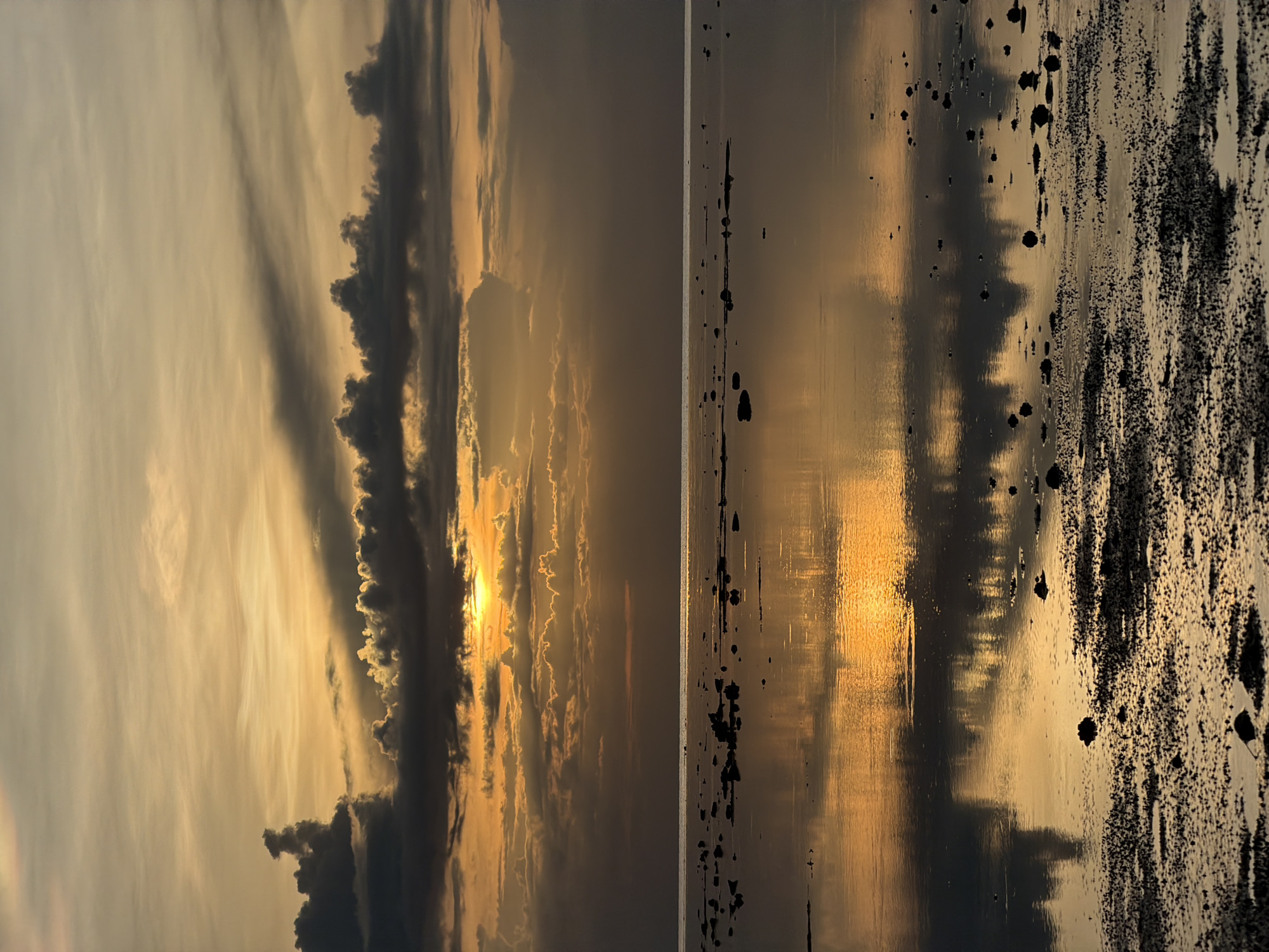 This image depicts a serene beach scene at sunset in Baan Plai Laem, Thailand. The sky is a canvas of warm hues, with golden sunlight breaking through dramatic, dark clouds. The sun itself is partially obscured, casting a vibrant reflection on the calm water. The foreground is a shallow, rocky shoreline, with small pebbles and debris scattered across the wet sand, also reflecting the colors of the sky. The water extends to the horizon, where it meets the cloudy sky. No people are visible in the frame.