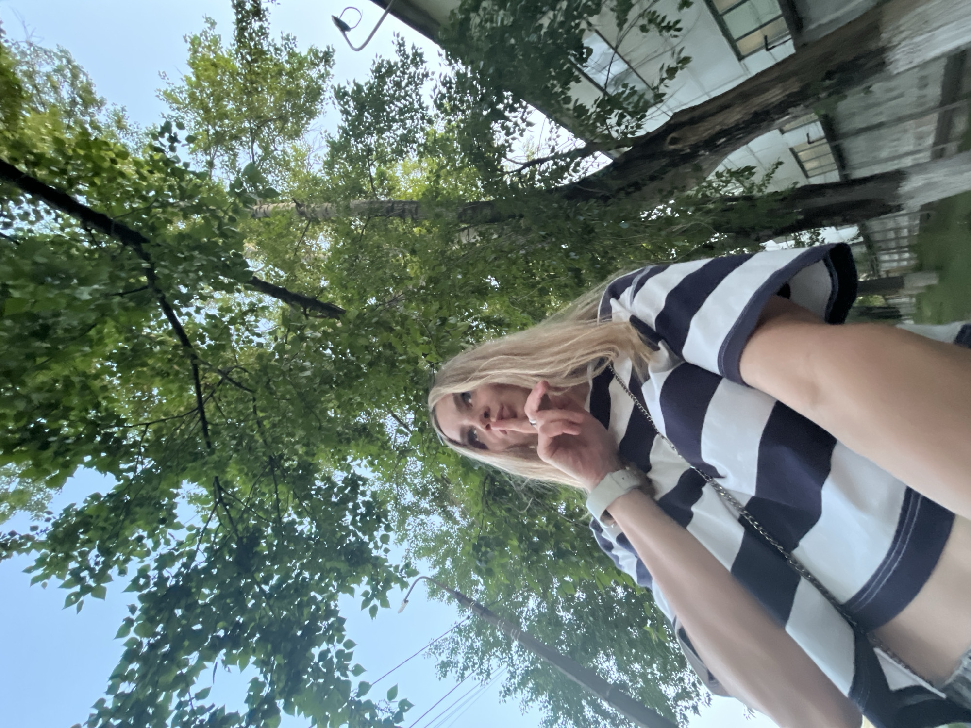 The image appears to be a selfie taken by a fair-skinned young woman with long blonde hair. She is wearing a navy and white striped top. She is holding a finger to her lips in a "shush" gesture. The background consists of green trees and sky, and a building. The photograph was taken in Chaikovsky, Russia.