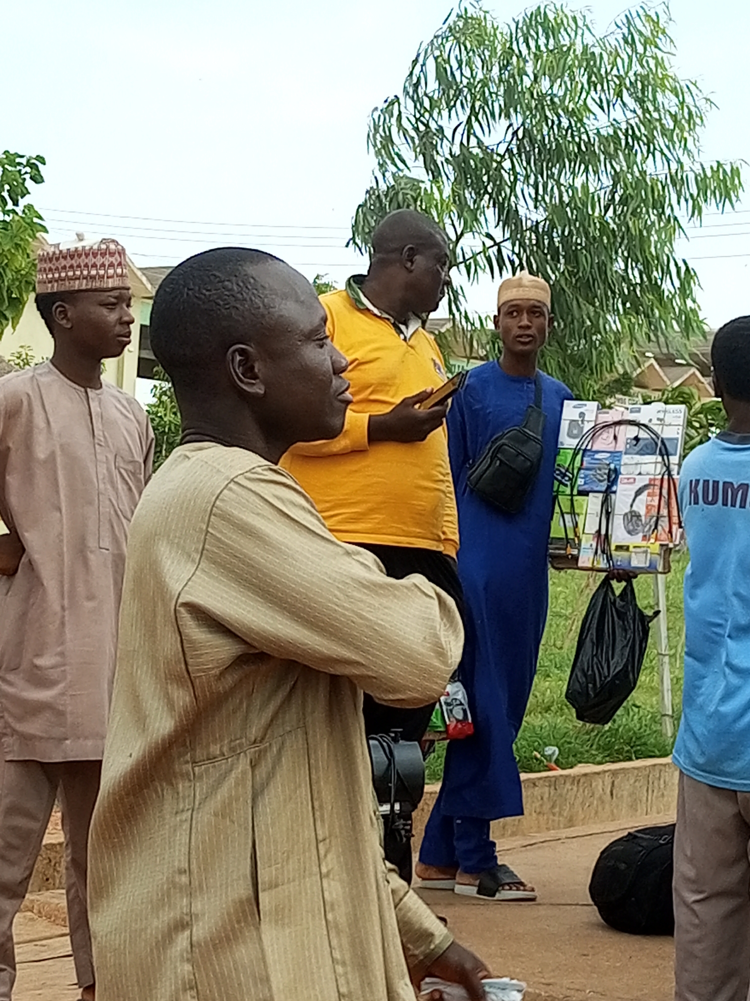 In the city of Gombe, Nigeria, a group of four men are gathered outdoors. The man in the foreground, on the left, is wearing a light brown striped garment and a cap. To his left, another man in a light brown outfit and a patterned cap stands partially visible. In the center of the image, a man in a bright yellow polo shirt holds a mobile phone. Beside him, a man in a blue robe is holding a tray of electronic items, possibly mobile phone accessories, and has a black bag slung across his chest. To the far right, a fourth man, wearing a blue t-shirt with the word "KUMO" on it, is also visible. The background features a large tree with green leaves and some buildings. The ground is paved.