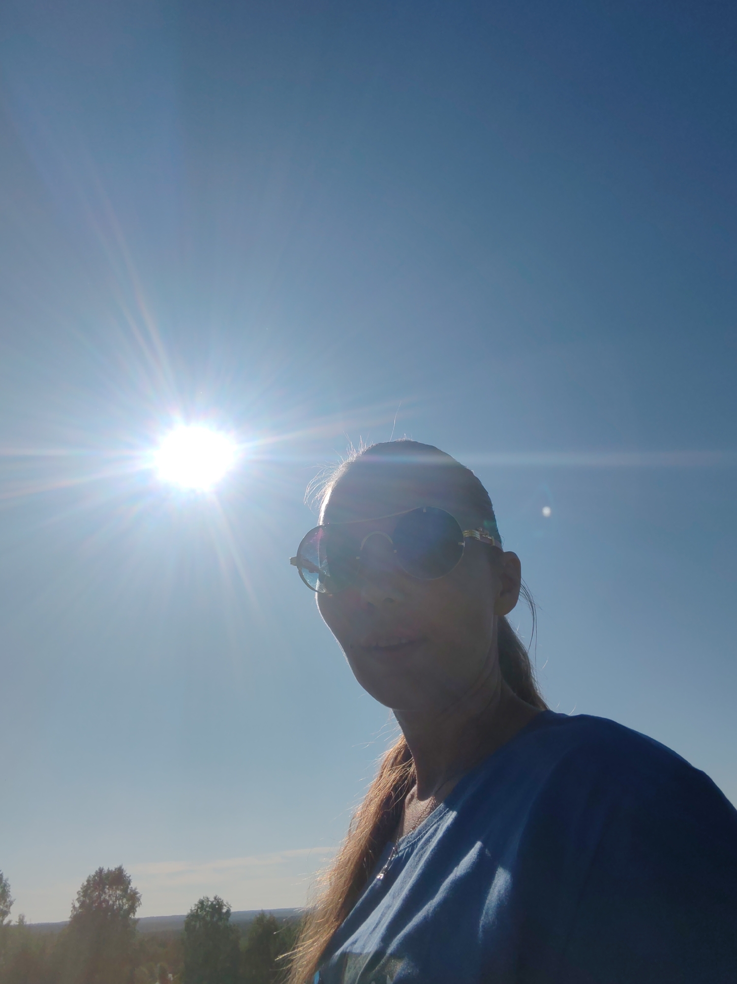 A woman is pictured from the chest up, outdoors, with a bright sun and a clear blue sky dominating the background. She is wearing large sunglasses with gold trim and a blue t-shirt. Her long, light brown hair is pulled back and cascades over her shoulder. The sun is positioned to the left of the frame, casting a strong glare and lens flare across the image. In the distance, a treeline is visible below a faint horizon. The image appears to have been taken in a rural or suburban outdoor setting.