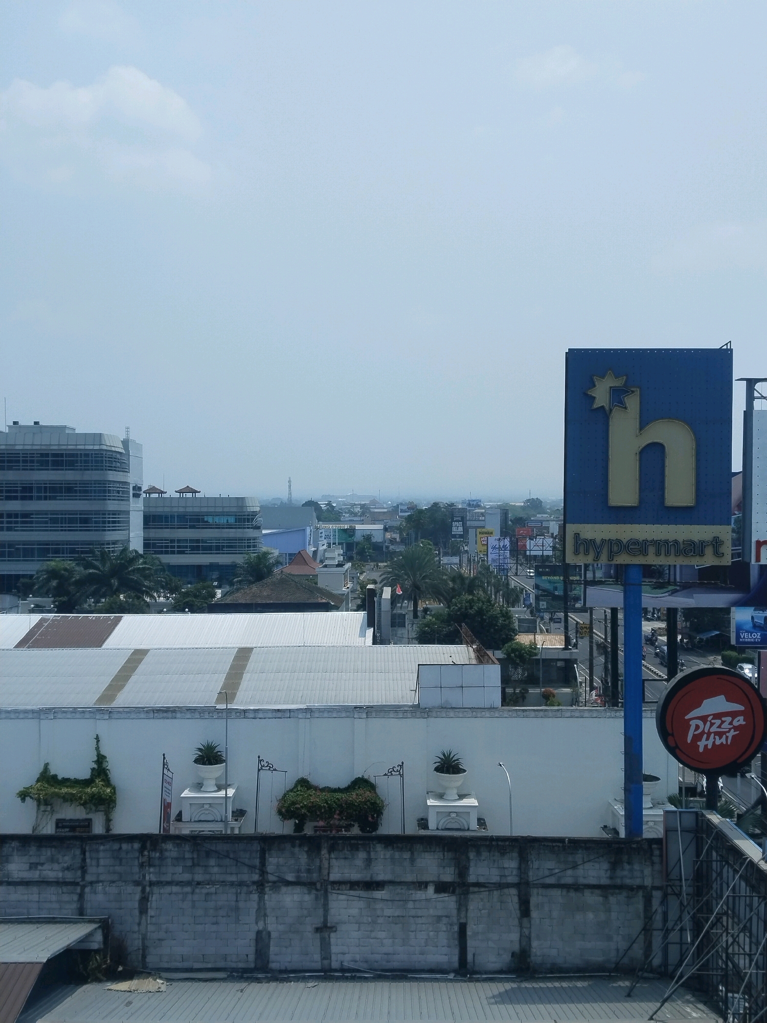 The image is a daytime, eye-level shot looking over a cityscape in Sinduadi, Indonesia. The sky is overcast with light blue tones. In the foreground, a gray concrete wall runs horizontally across the bottom of the frame, with a pitched metal roof visible below it on the left. Above the wall, a white building with a large, flat white roof is visible, featuring two white decorative urns with small plants. To the left of these urns, green vines climb a structure. Further back, a line of buildings of varying heights and architectural styles are seen. Prominently displayed on the right is a large, blue sign for "Hypermart," featuring a golden stylized letter "h" and a starburst. To the right of the Hypermart sign, a red circular sign for "Pizza Hut" is visible. Beyond these signs, a street with traffic and other billboards can be seen, leading into the distance where the city fades into a hazy horizon. Palm trees are scattered throughout the urban landscape.