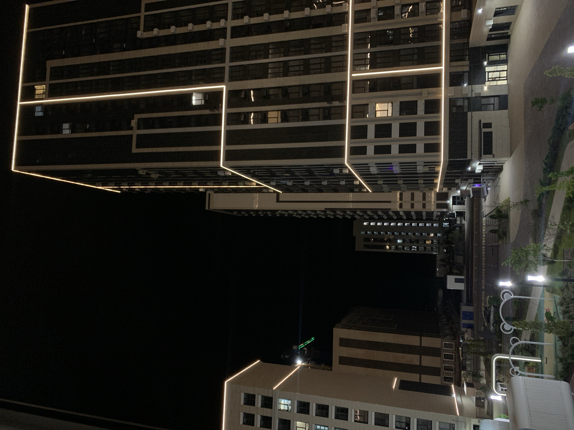 The image captures a nighttime view of a modern residential complex in Stavropol, Russia. The scene is dominated by several tall, contemporary buildings featuring dark facades accented with illuminated strips of light, highlighting architectural lines. One prominent building on the right is a high-rise with a grid-like pattern of windows and vertical beige and dark gray panels, outlined by warm, white LED lighting along its edges and dividing elements. To the left, another lighter-colored building stands, also with illuminated horizontal bands and numerous windows. In the middle ground, a crane, likely for ongoing construction, is visible with a green light on its jib. The foreground shows a playground area with swings and decorative illuminated posts, suggesting a family-friendly environment. The ground is paved, and there are areas of landscaping with trees and grass. The overall impression is one of urban development at night, with artificial lighting playing a significant role in the visual aesthetic.
