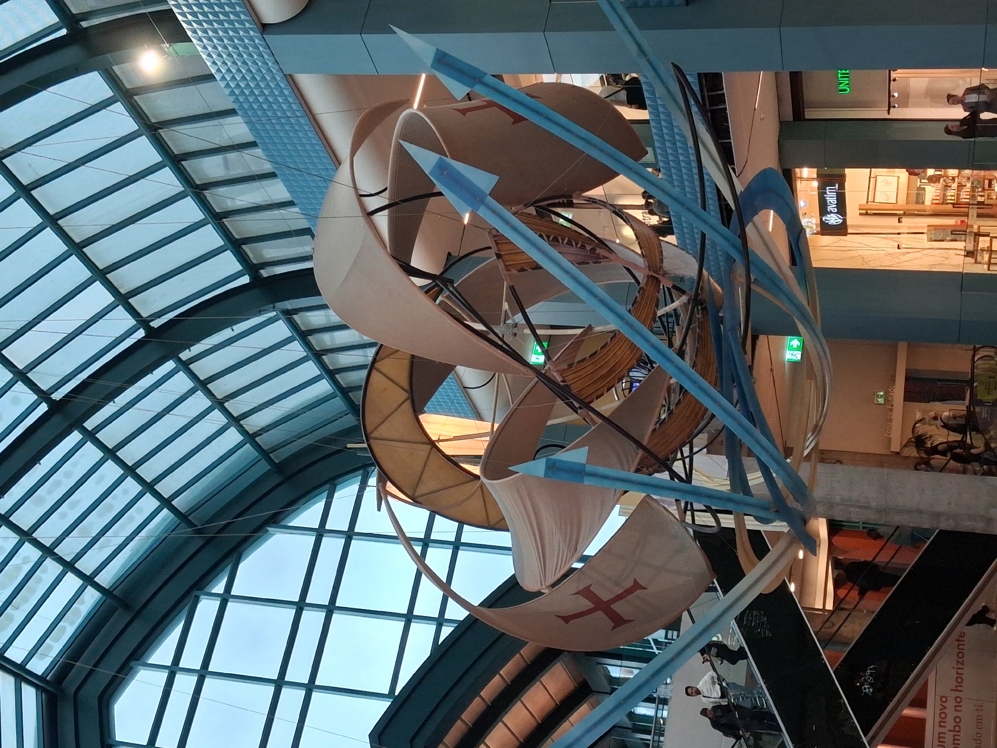 The image captures a view from below of a complex architectural sculpture in what appears to be a shopping mall or large public space. The sculpture incorporates stylized sails, some bearing a red cross design, intertwined with blue tubular structures and geometric forms, creating a dynamic visual element in the open space. The setting is enclosed by a large, arched glass ceiling, allowing ample natural light to enter. In the background, glimpses of retail spaces, signage, and people walking on upper levels can be seen, indicating the commercial nature of the location. The overall impression is one of modernity, with the sculpture serving as a focal point within the architectural context.
Based on the signage and architectural style, it appears to be a mall in Lisbon, Portugal, due to the presence of the "avatin" store logo, common in Portugal.