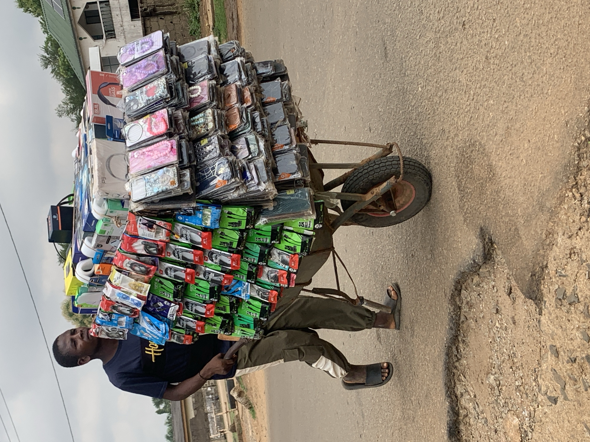 The image captures a man pushing a heavily laden handcart along a paved road in Maigizo, Nigeria. The handcart, constructed from metal with a single wheel, is piled high with various electronic accessories.
The merchandise predominantly consists of numerous phone cases, arranged in stacks. These cases display a variety of colorful and patterned designs, some featuring decorative elements like beads or intricate floral patterns. Below the phone cases, many packaged earphones and other accessories are visible. Several green packages for earphones clearly show text such as "2A FAST" and "M11". A box for headphones, labeled "SUPER BASS," is also part of the load.
The man, appearing to push or pull the cart, is dressed in a dark t-shirt with a yellow logo or design on the front, dark green cargo pants with white stripes along the sides, and black flip-flops. His expression is neutral, focused on transporting the goods.
The scene takes place outdoors during the daytime, under an overcast sky. The road surface is asphalt, showing signs of wear with visible cracks and rough patches. To the side of the road, there is exposed dirt and gravel. In the background, a building with a distinctive green roof and solar panels is visible, along with some trees, suggesting a semi-urban or rural setting. The image depicts a common street vending activity.