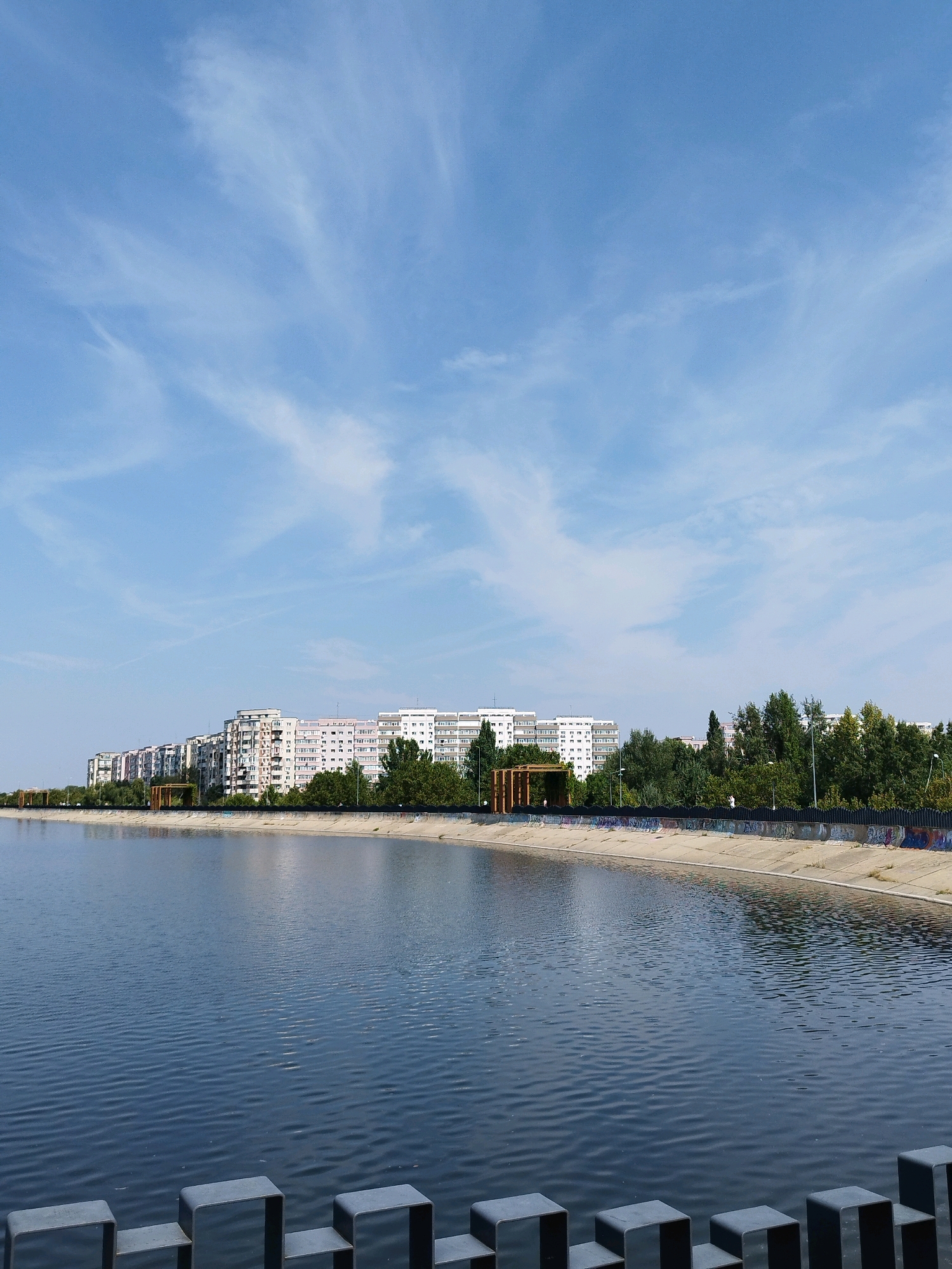 This is a photograph of the Vacaresti Lake Natural Park in Bucharest, Romania. The image features a wide expanse of dark, rippling water in the foreground, bordered by a concrete embankment. Along the embankment, there are sections of graffiti and a low black fence with square posts. Behind the embankment, a line of trees stands, with several tall, multi-story apartment buildings visible above them. The sky is a clear blue with wispy white clouds. There are no people or animals readily visible in the scene. The overall impression is one of an urban park environment, with the lake and greenery providing a contrast to the surrounding residential buildings.